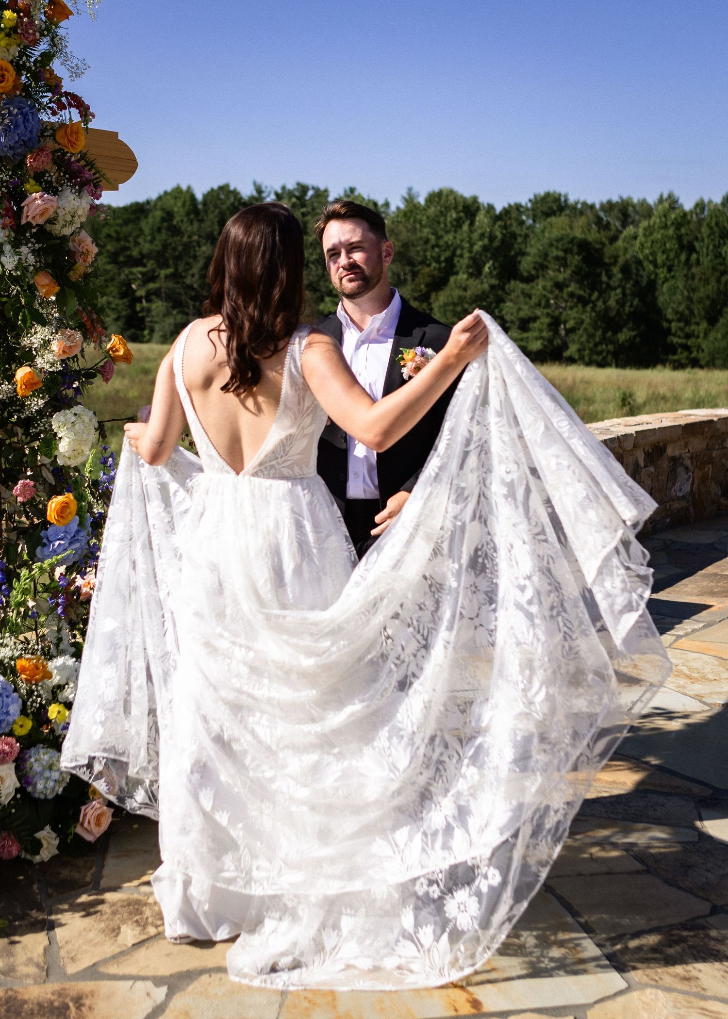 Groom lifting bride's lace wedding dress train outdoors near floral arch on sunny day.