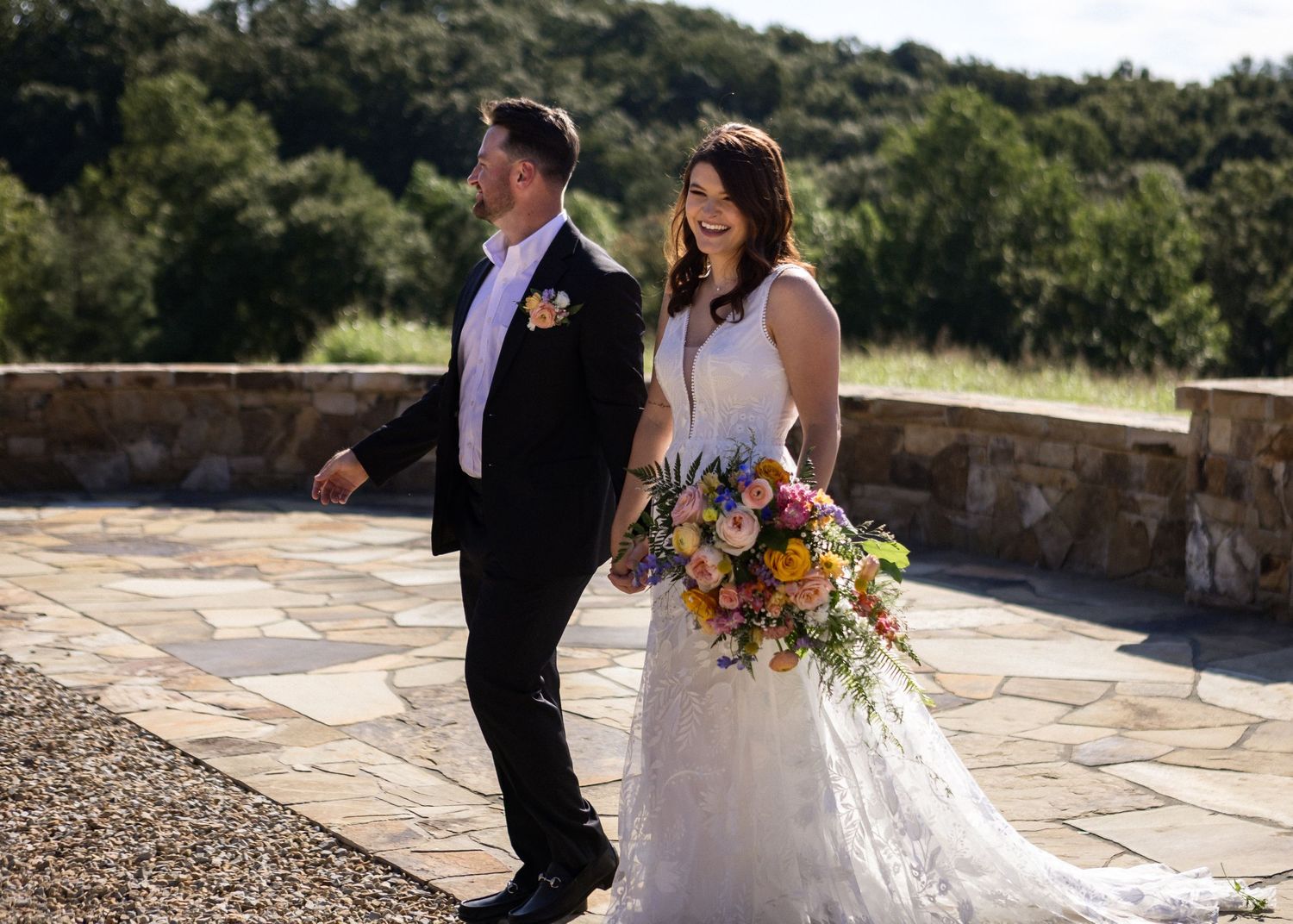 Smiling bride in lace gown holds colorful bouquet while walking with groom on stone patio with scenic hills.