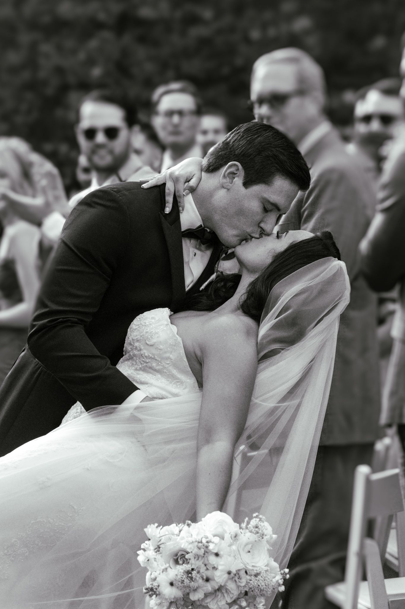 Bride and groom share a romantic dip kiss at outdoor wedding ceremony with guests watching in background.