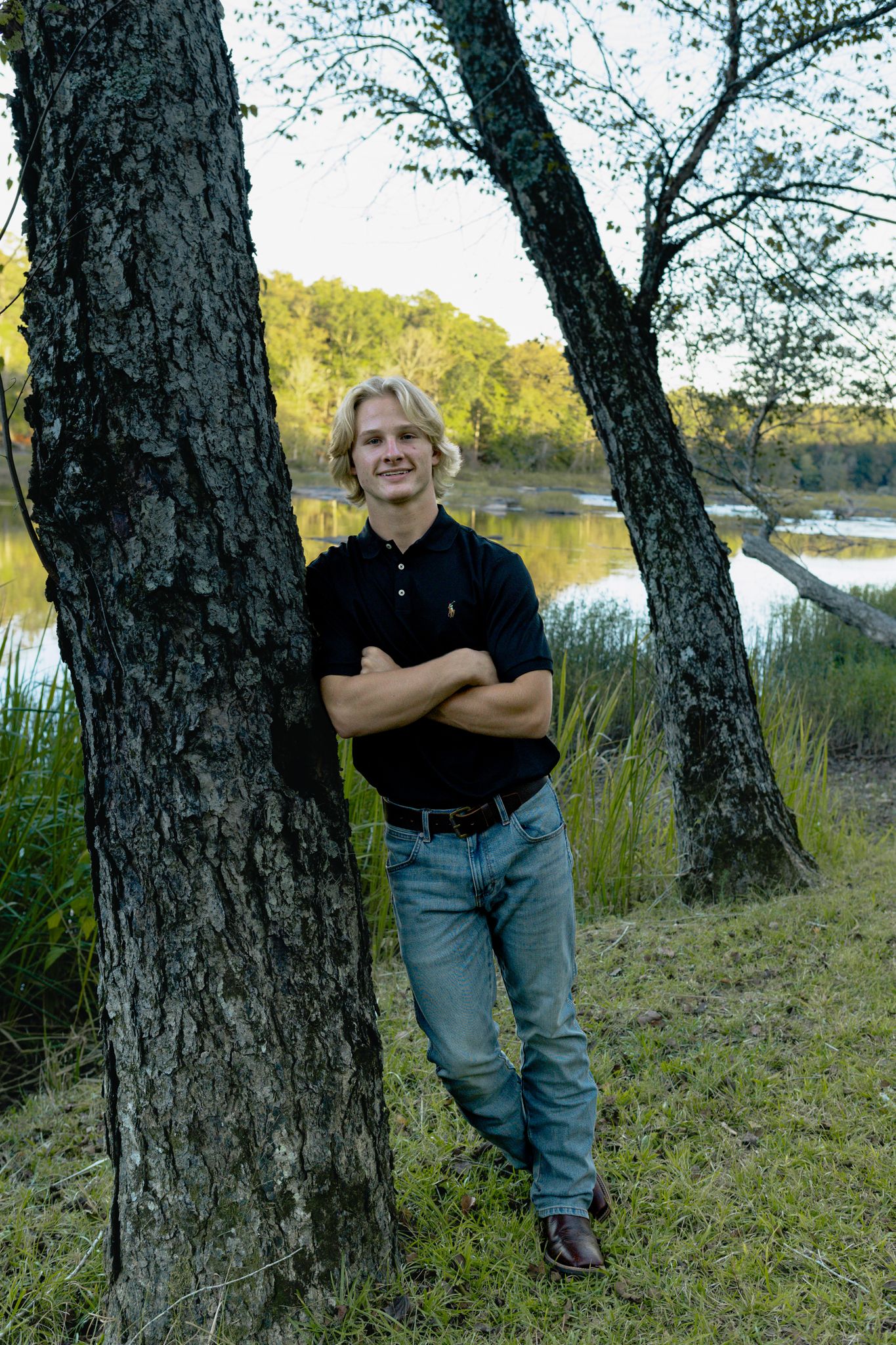 Young man with blond hair leans against a tree with arms crossed, smiling near a serene lake backdrop.