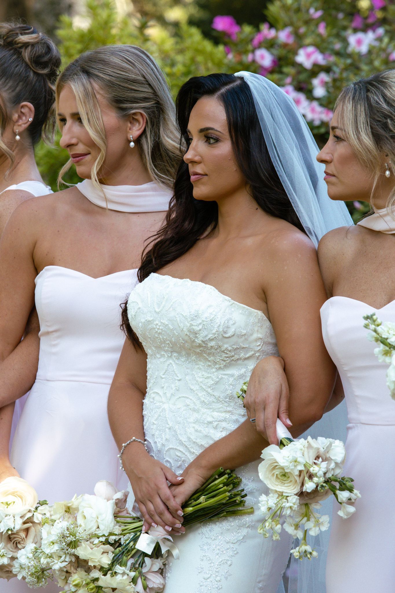 Bride in strapless lace gown with veil poses with bridesmaids in white dresses holding bouquets.