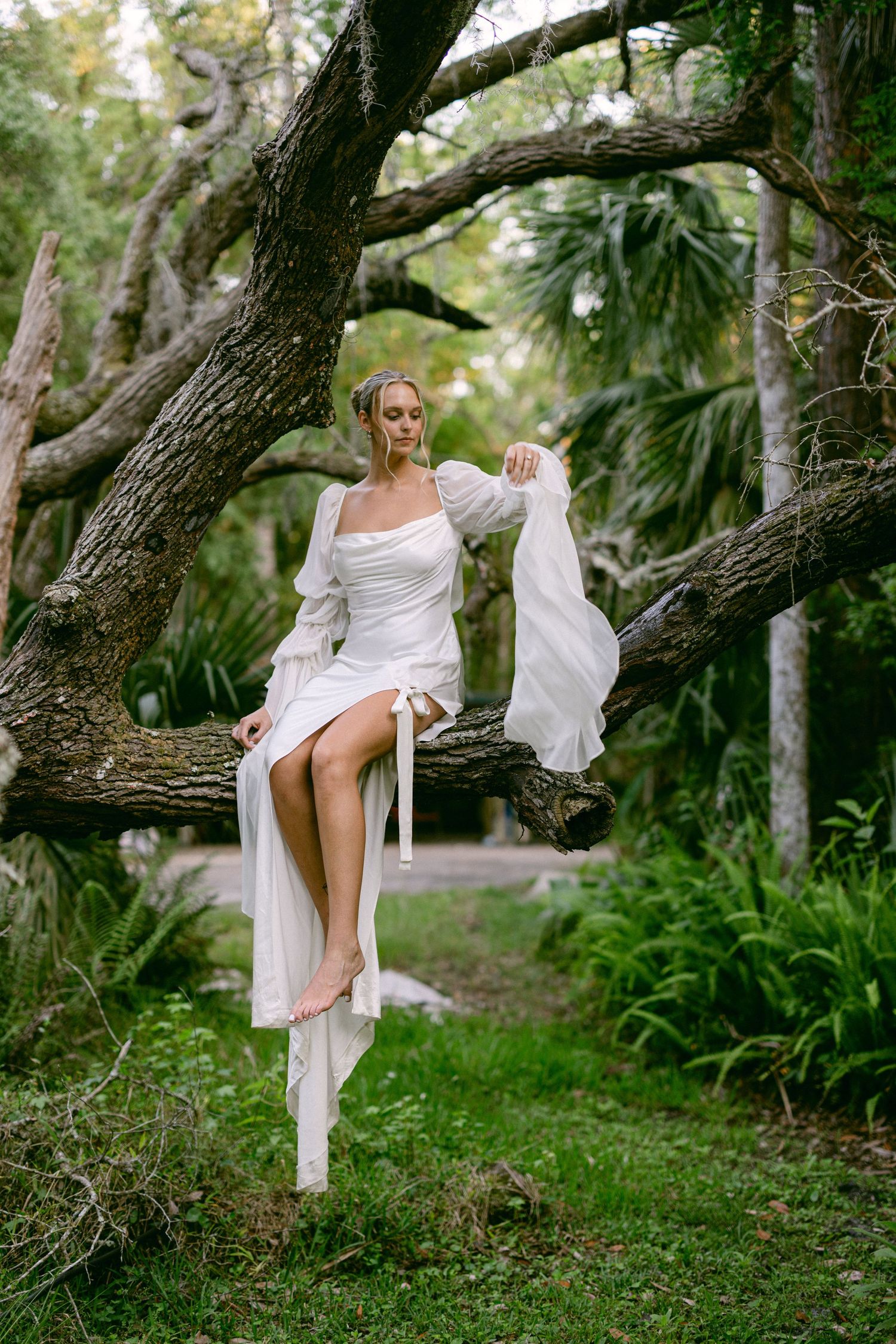 Bride in white square-neck gown with billowy sleeves sitting barefoot on a large tree branch in lush garden.