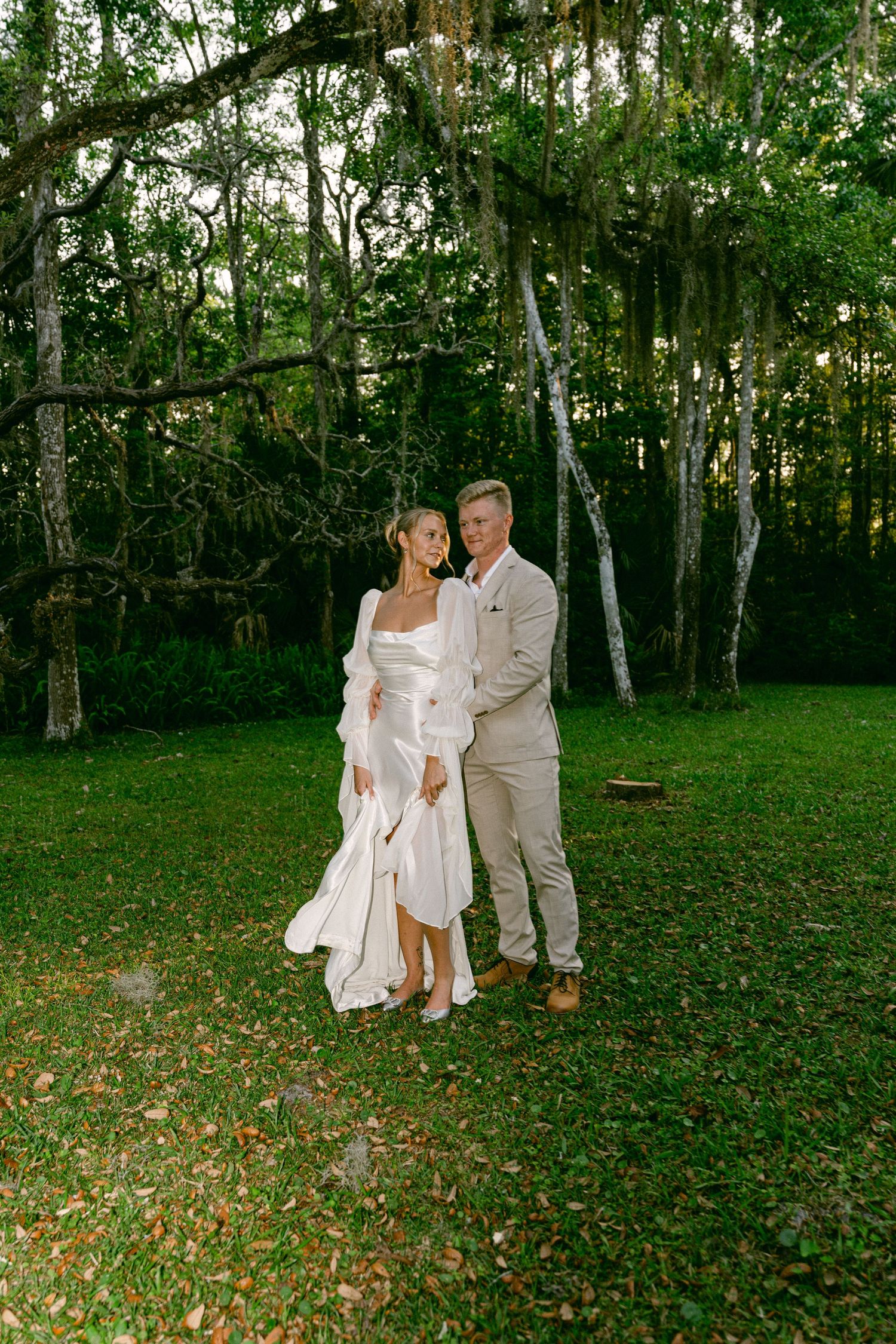 Smiling bride and groom walking hand-in-hand under Spanish moss-covered oak trees at sunset.