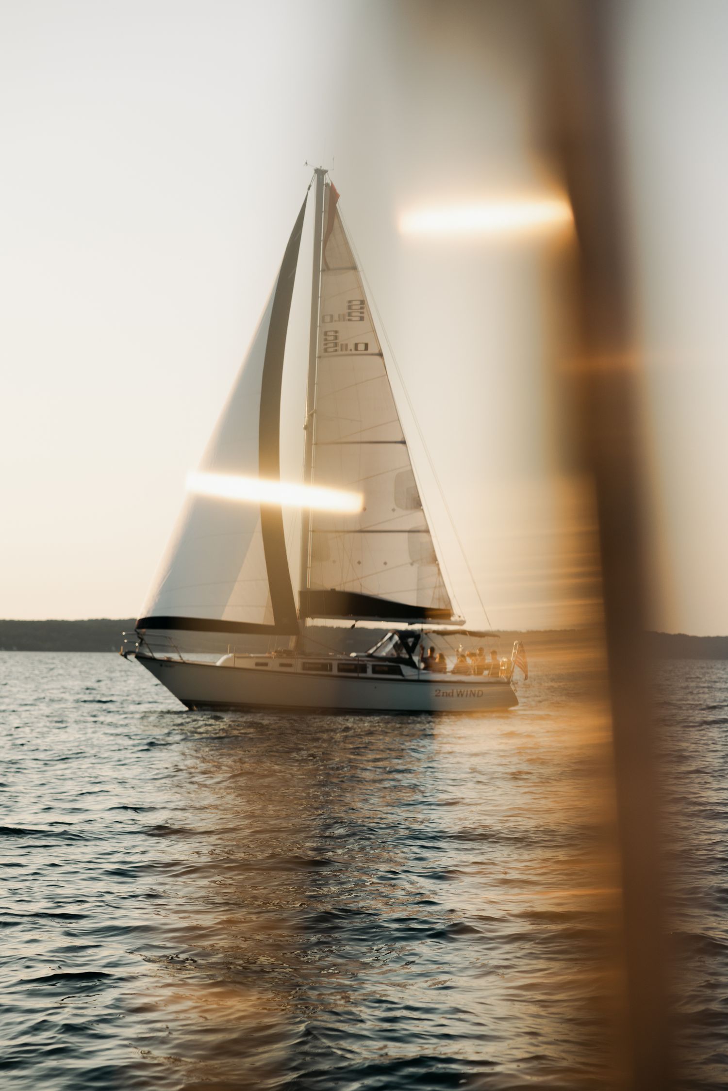 Sailboat with white sails gliding on calm water at golden sunset, viewed through soft bokeh foreground.