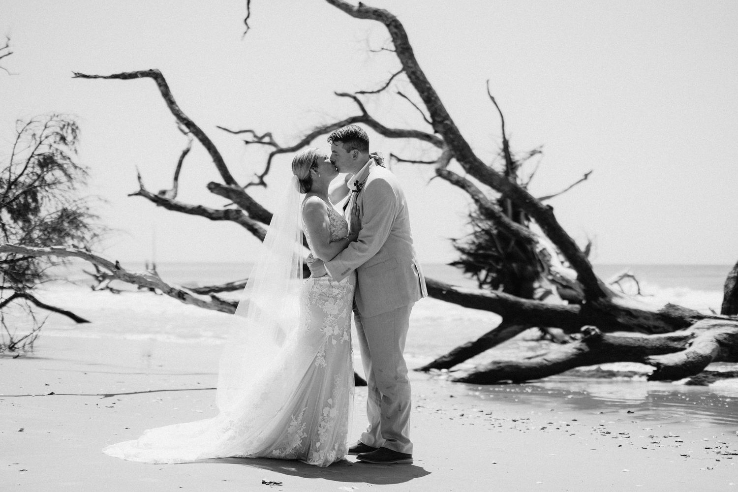 Black and white beach wedding photo of bride and groom kissing near driftwood by the ocean.