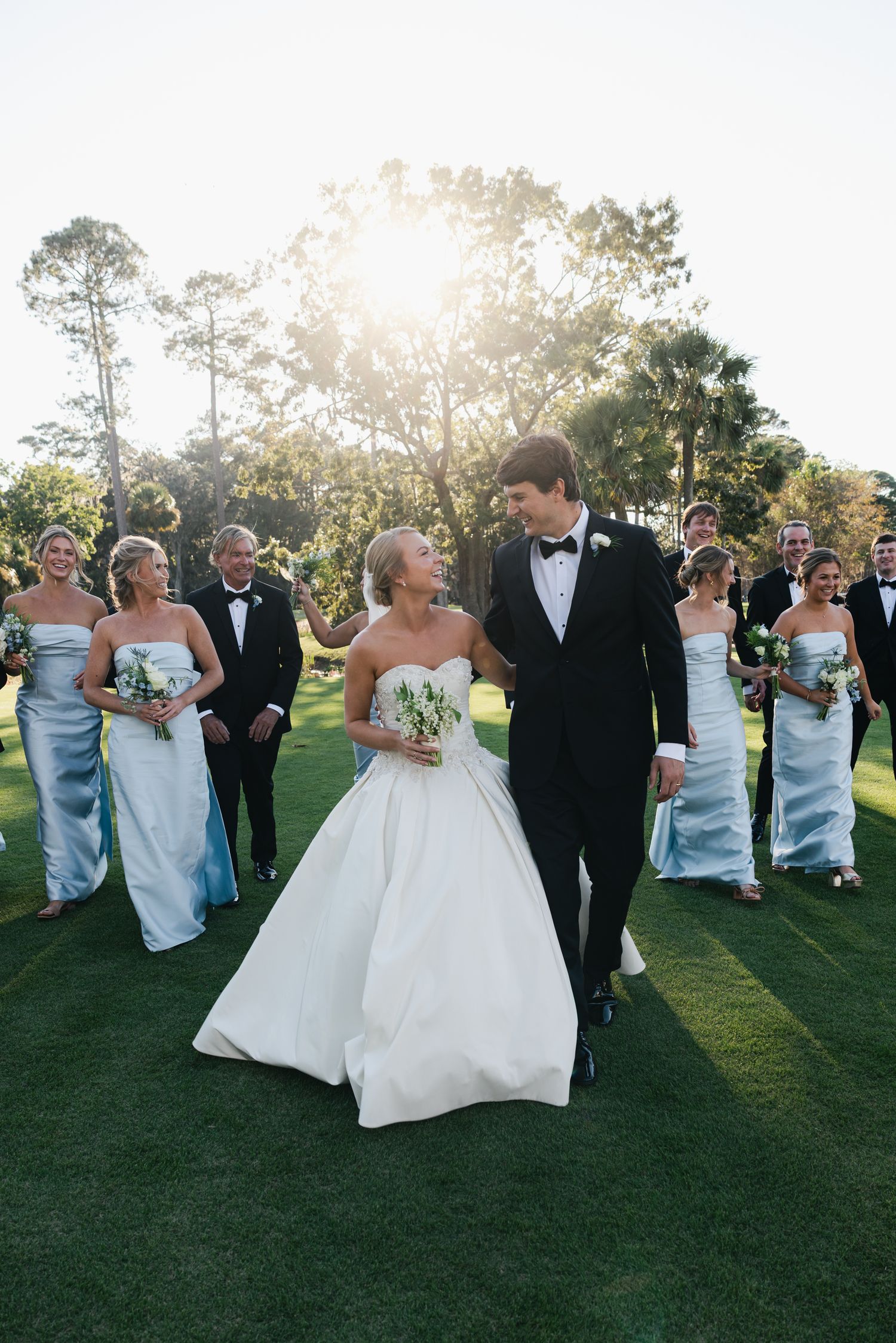 Bride and groom walk together on green lawn with wedding party in light blue gowns and tuxedos in golden sunlight.