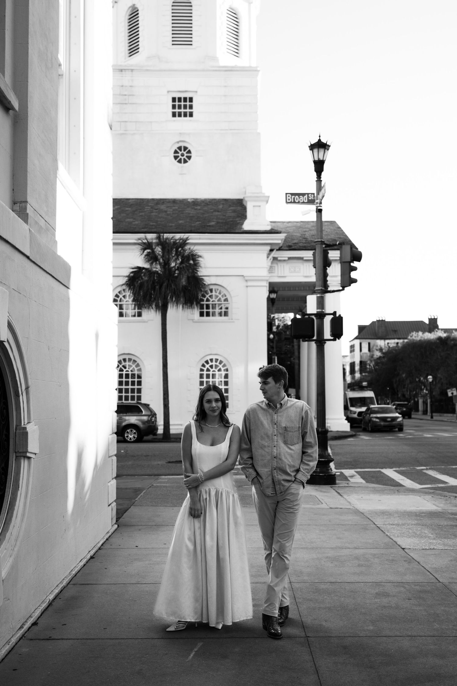 Black and white photo of a couple walking on a sidewalk in front of a historic white church in Charleston, SC.