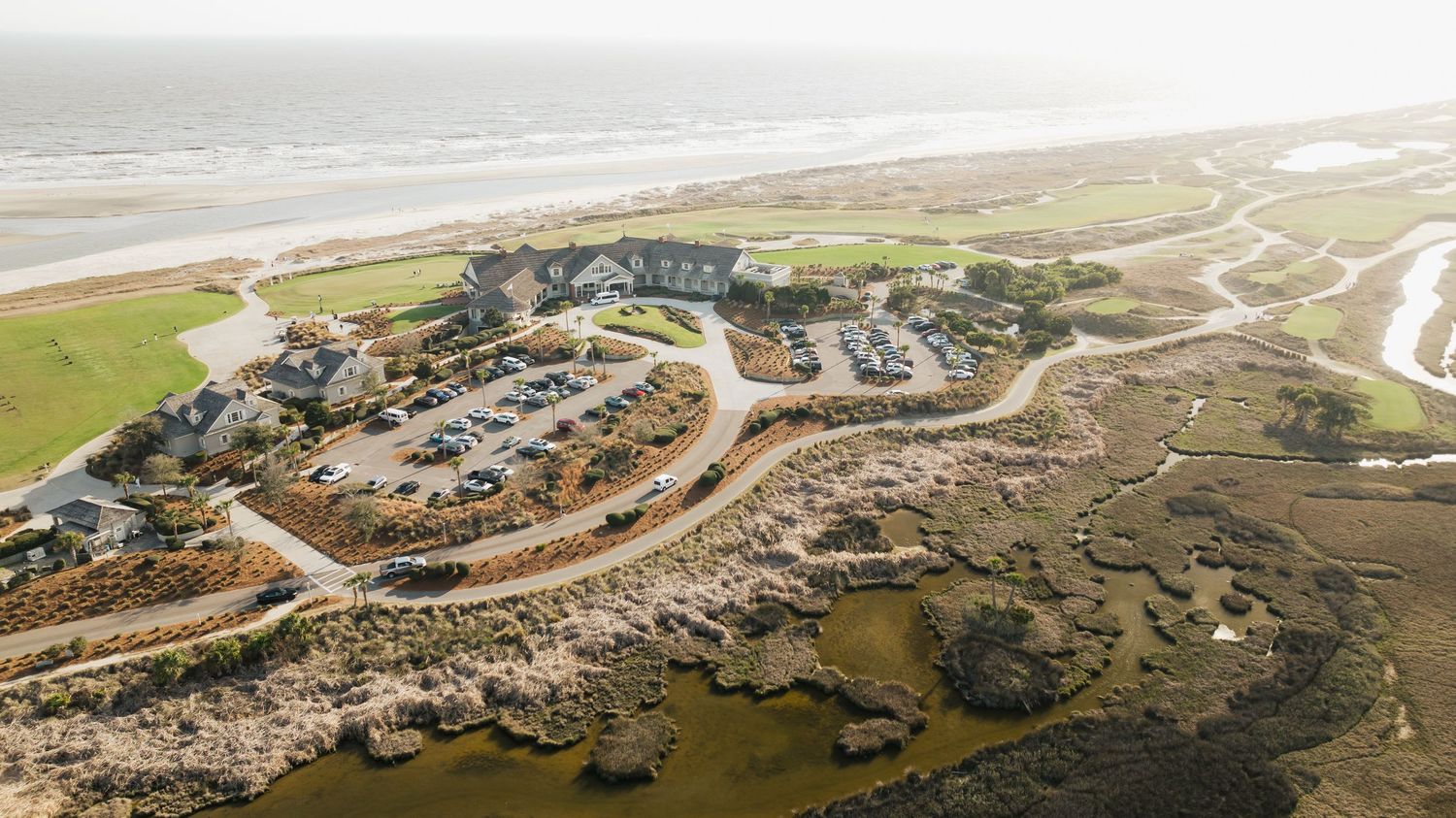 Aerial view of a coastal resort nestled among sand dunes, with ocean waves, green fairways, and winding paths visible.