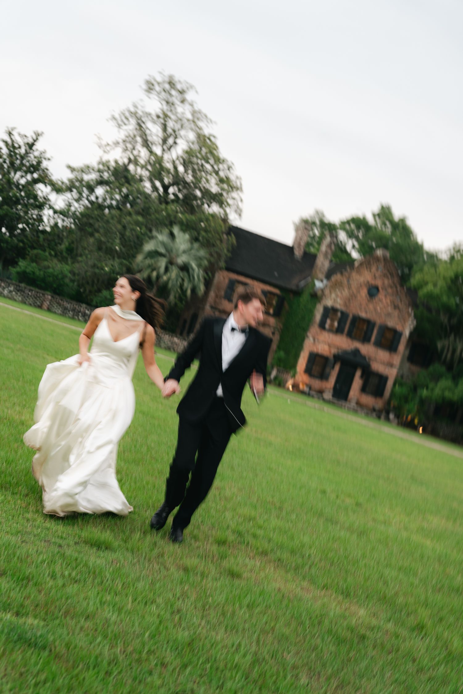 Joyful bride and groom running hand-in-hand across green lawn with historic brick manor in background.