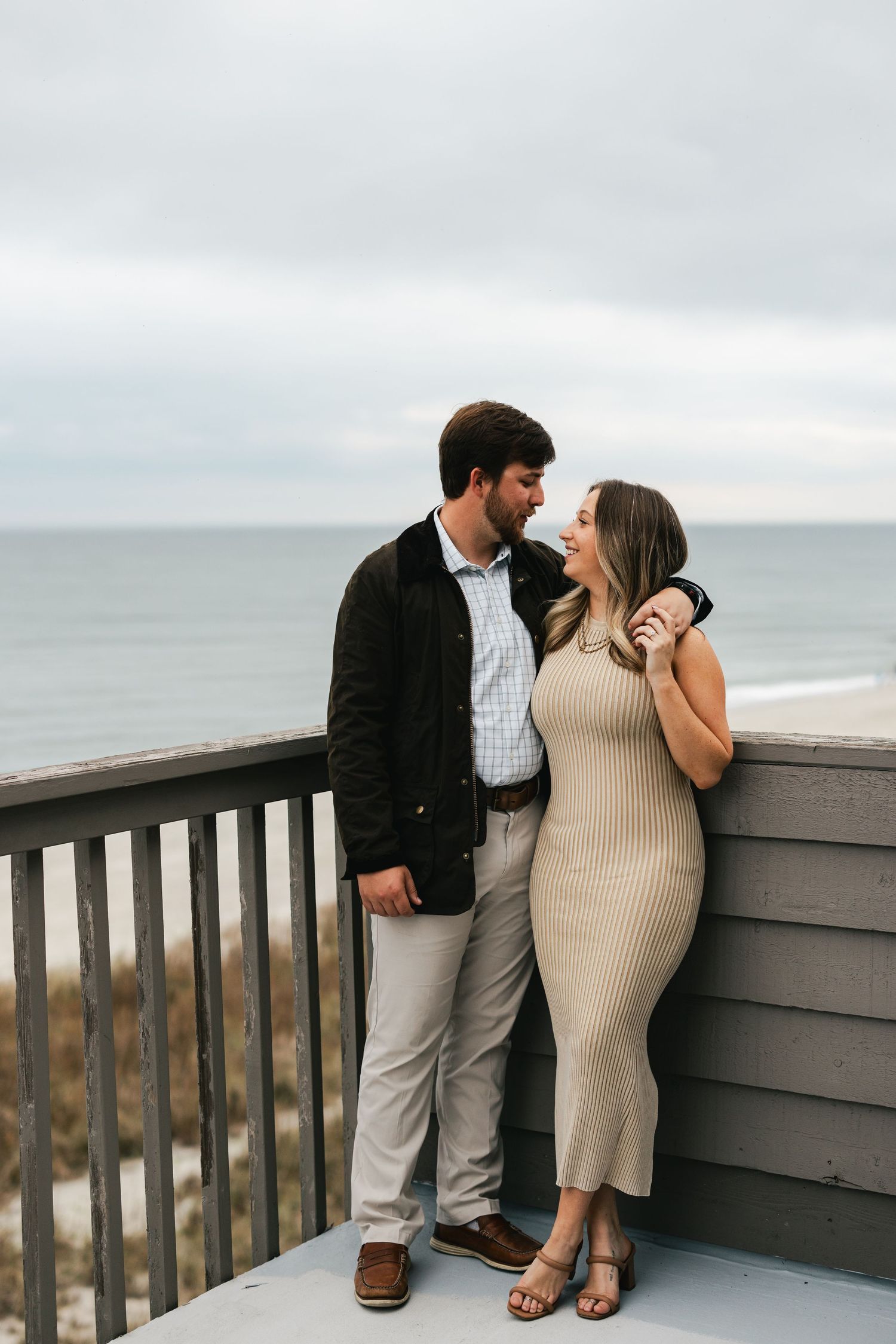 Couple sharing a romantic moment on a beachside deck, woman in beige ribbed dress, man in dark jacket, ocean backdrop.