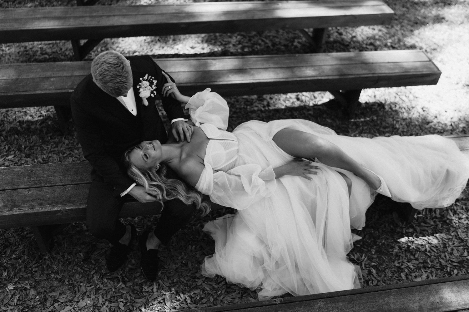 Black and white wedding photo of bride lying on groom's lap on a bench, holding flowers, in a romantic outdoor setting.