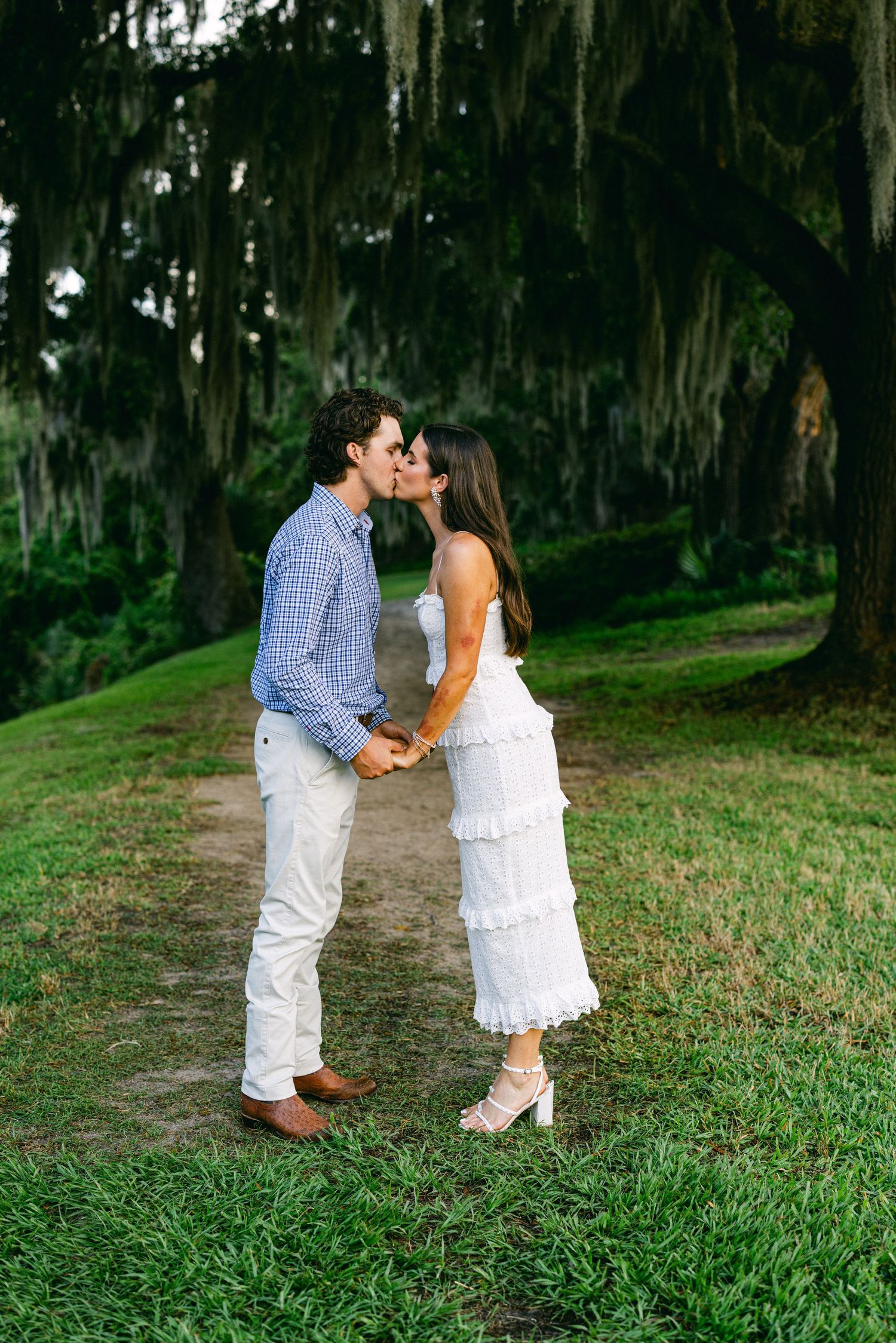 Couple sharing a romantic kiss while holding hands on a lush green path beneath Spanish moss-draped trees.