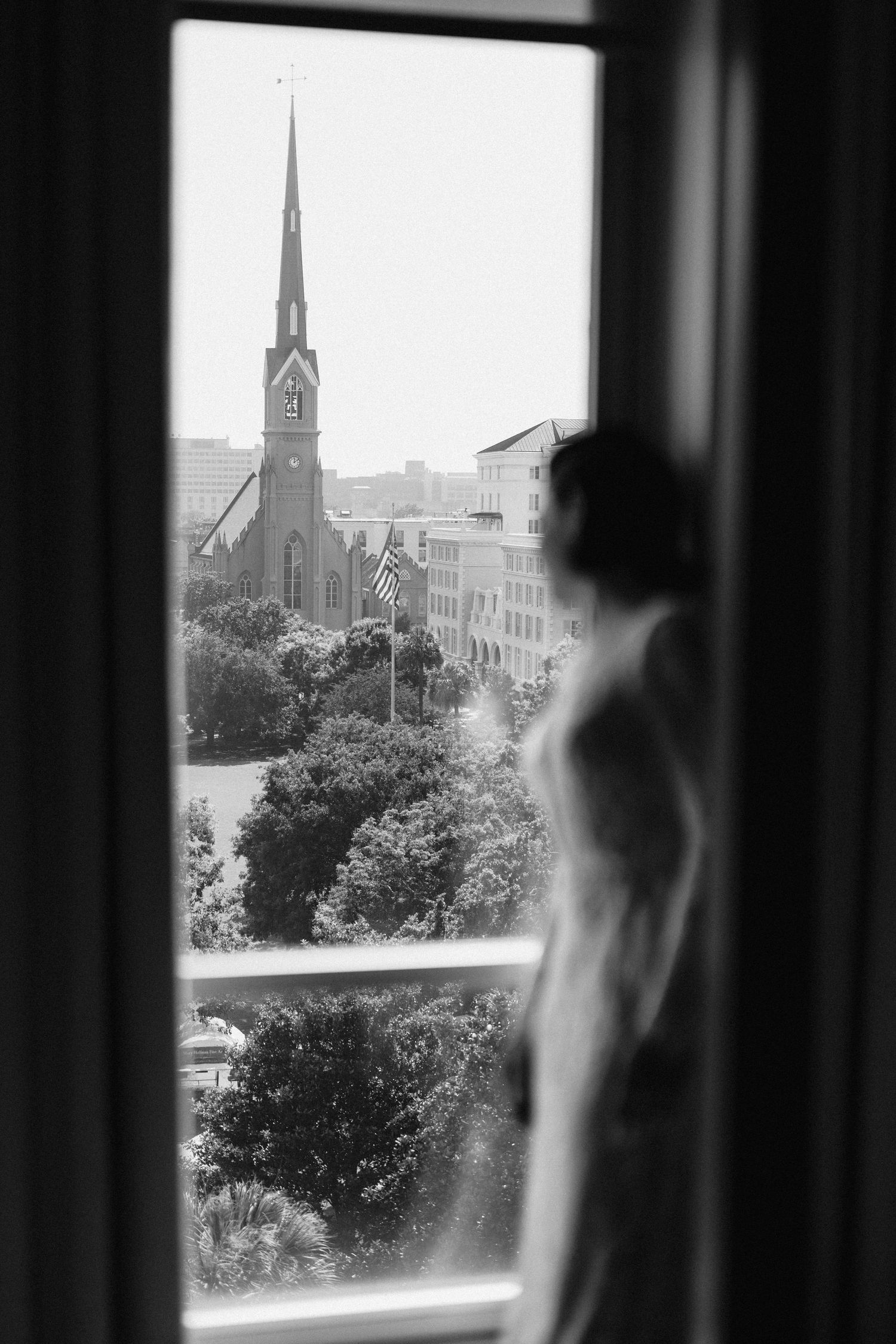 Black and white photo of a bride gazing out a window at a church steeple and city skyline.