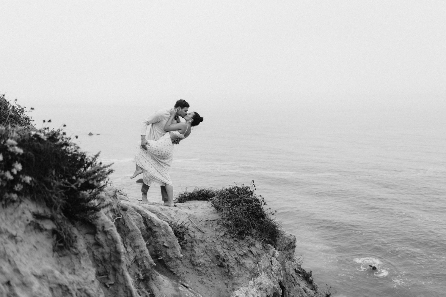 Black and white romantic photo of couple kissing on dramatic ocean cliff edge.