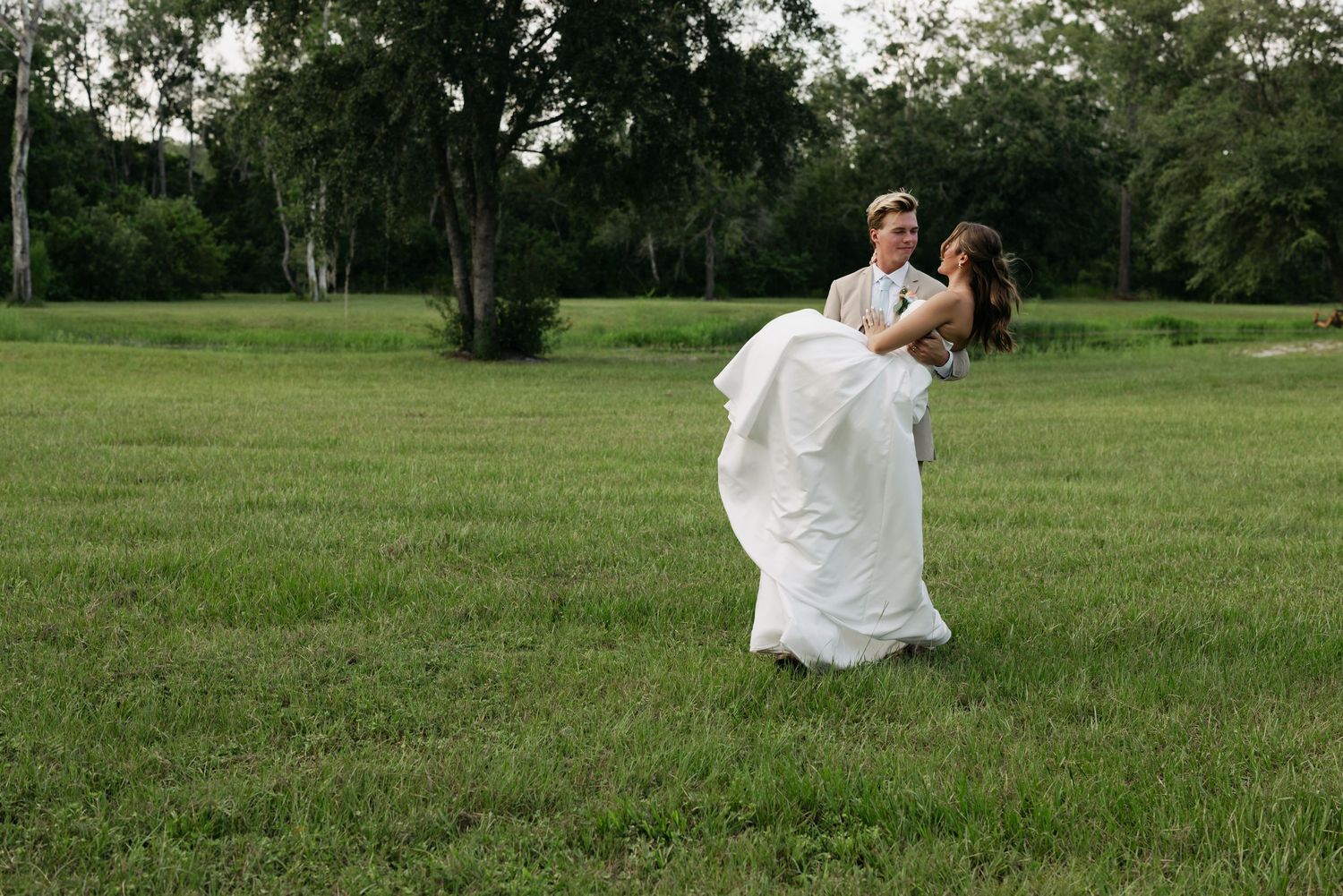 Groom lifting bride in white gown in lush green field with trees, romantic outdoor wedding photo.