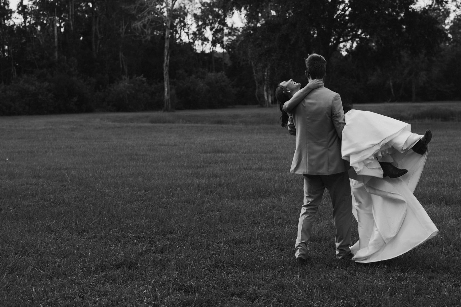 Black and white photo of bride and groom walking across lush green field, wedding dress flowing in the breeze.
