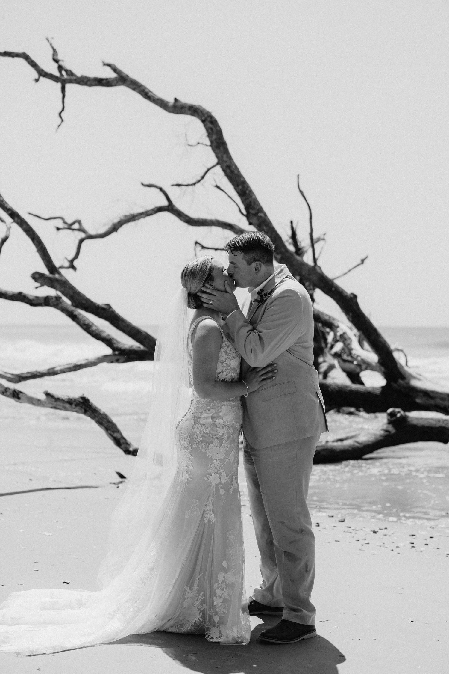 Black and white beach wedding photo of bride and groom sharing a kiss near driftwood on a sandy shore.