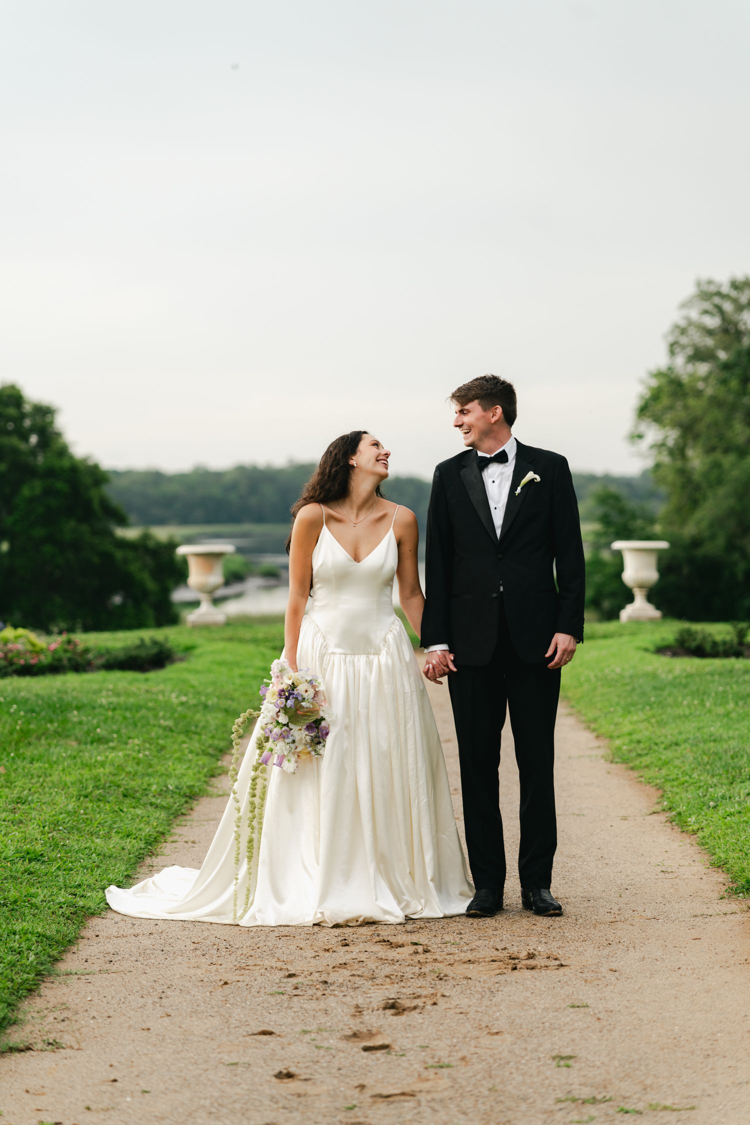 Bride and groom walking hand-in-hand on garden path, she in white gown with bouquet, he in black tuxedo.