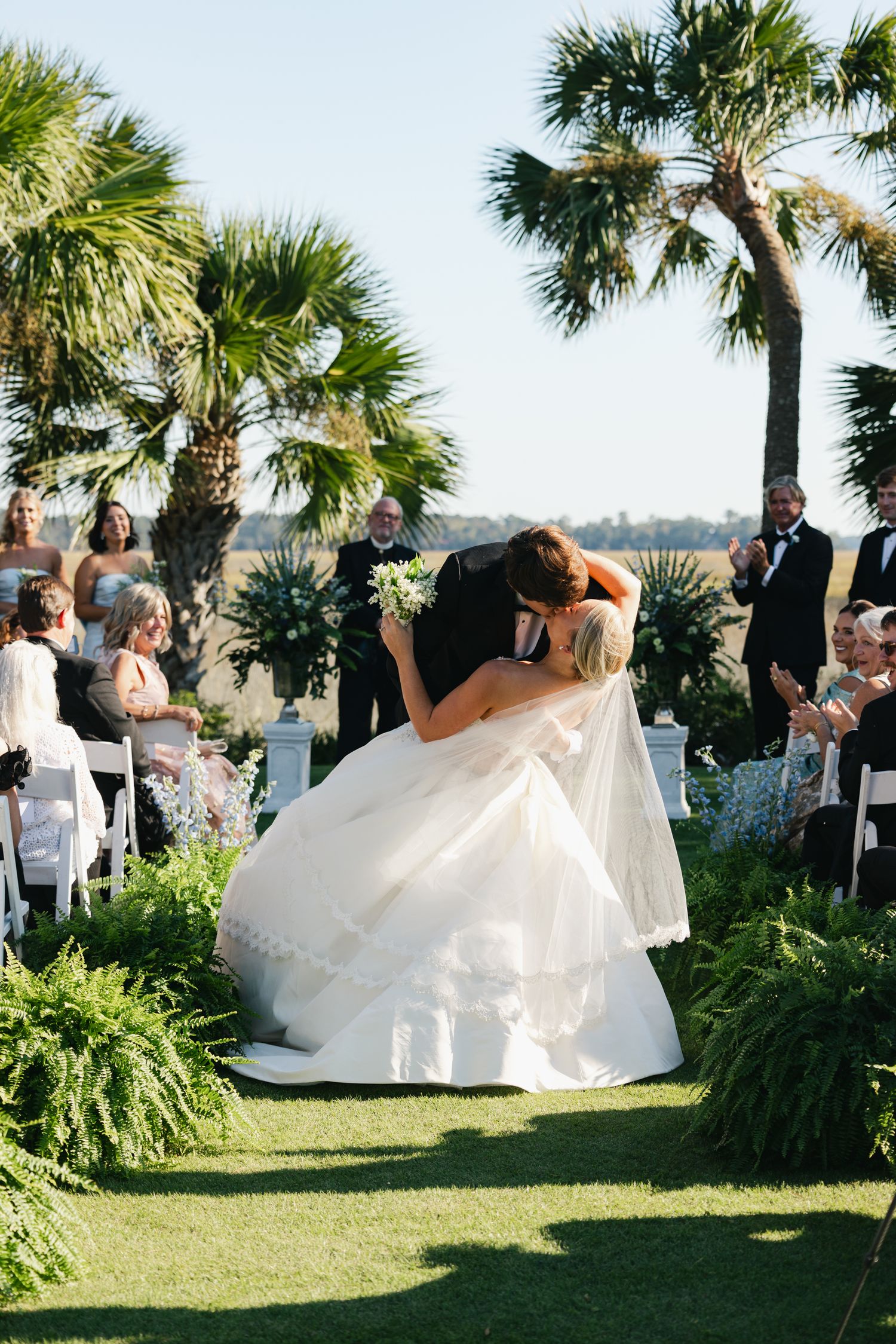 Bride and groom share a romantic dip kiss at outdoor wedding ceremony surrounded by palm trees and guests.