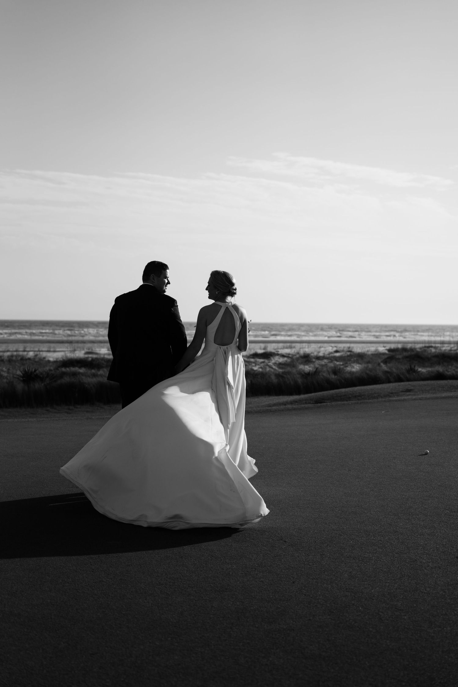 Black and white photo of a bride and groom walking hand in hand along a coastal road, wedding dress flowing in the wind.