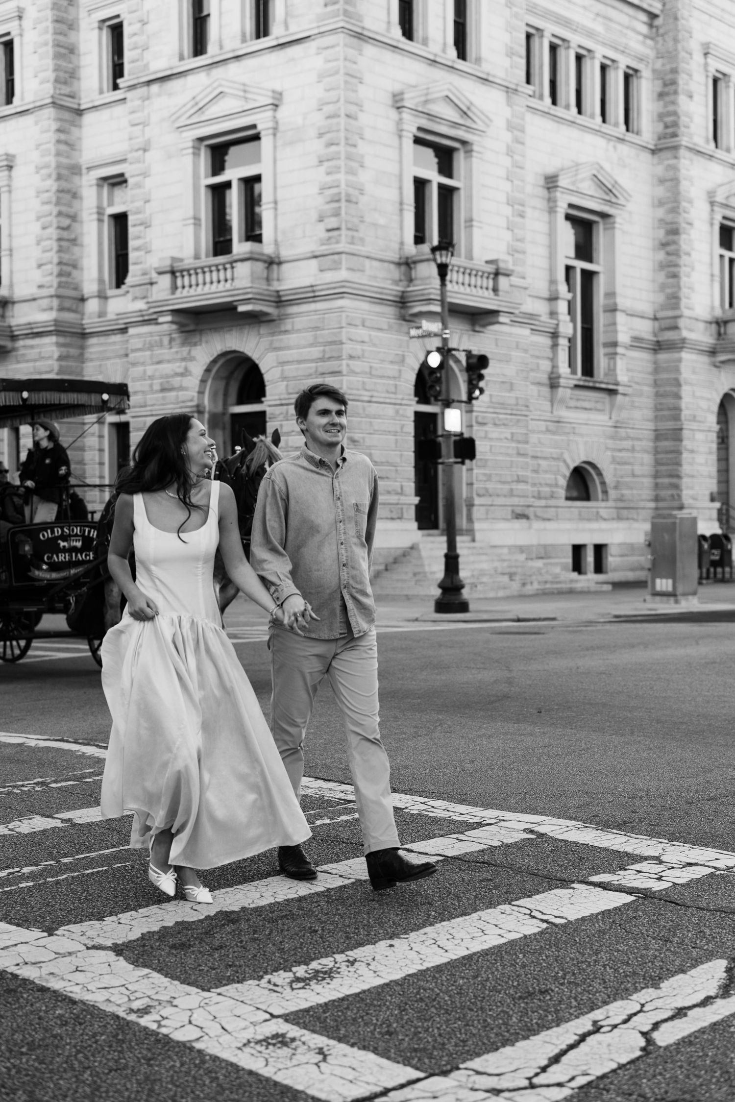 Couple holding hands crossing a street in black and white photo with elegant historic building backdrop.