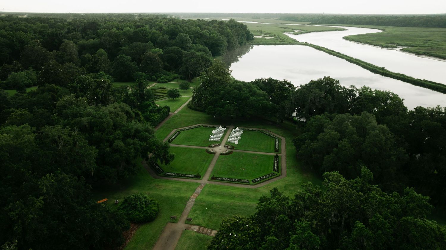 Aerial view of a historic plantation estate with manicured gardens and lawns bordered by lush trees and a winding river.