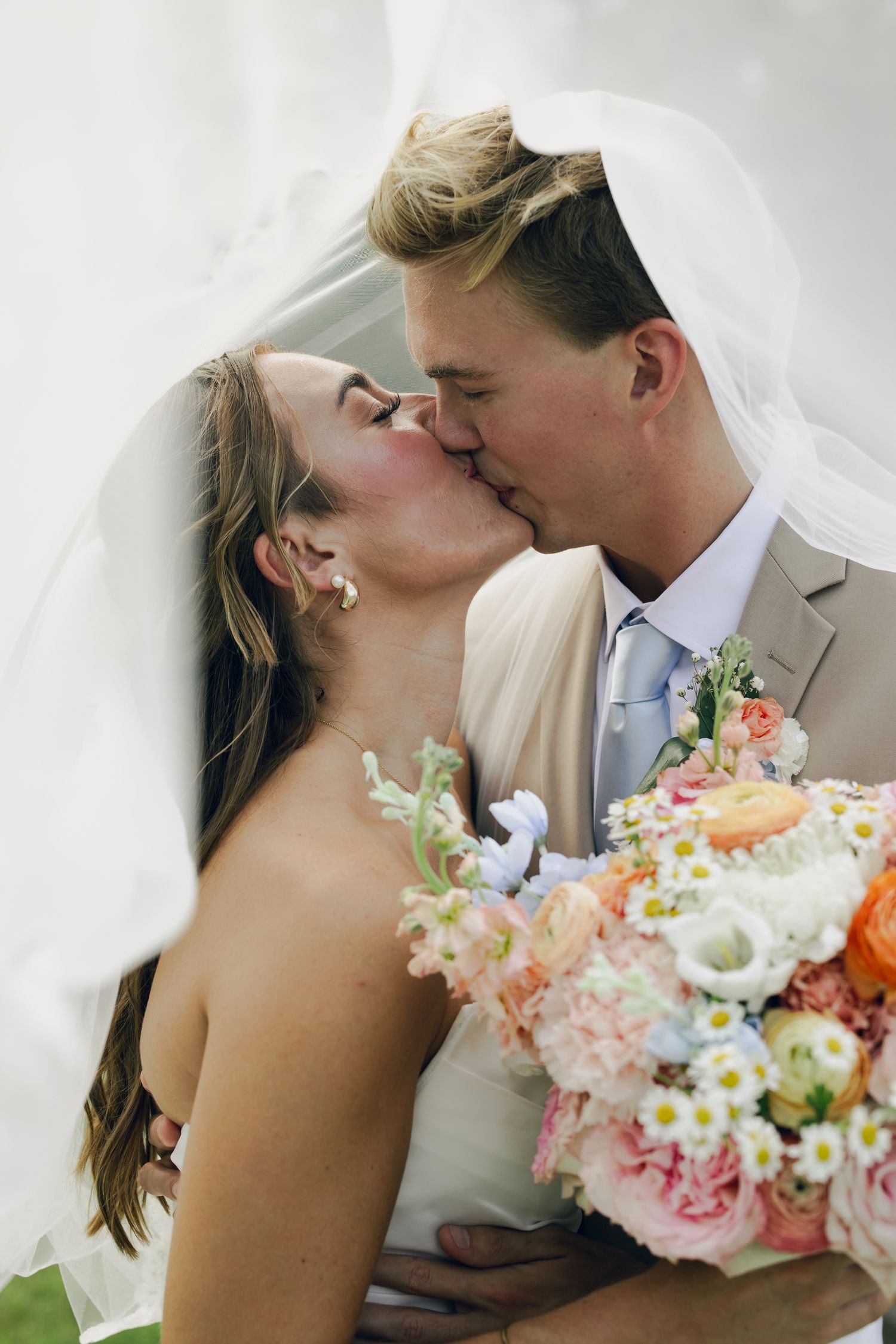 Bride and groom sharing a romantic kiss under a flowing veil, bride holding a pastel floral bouquet.