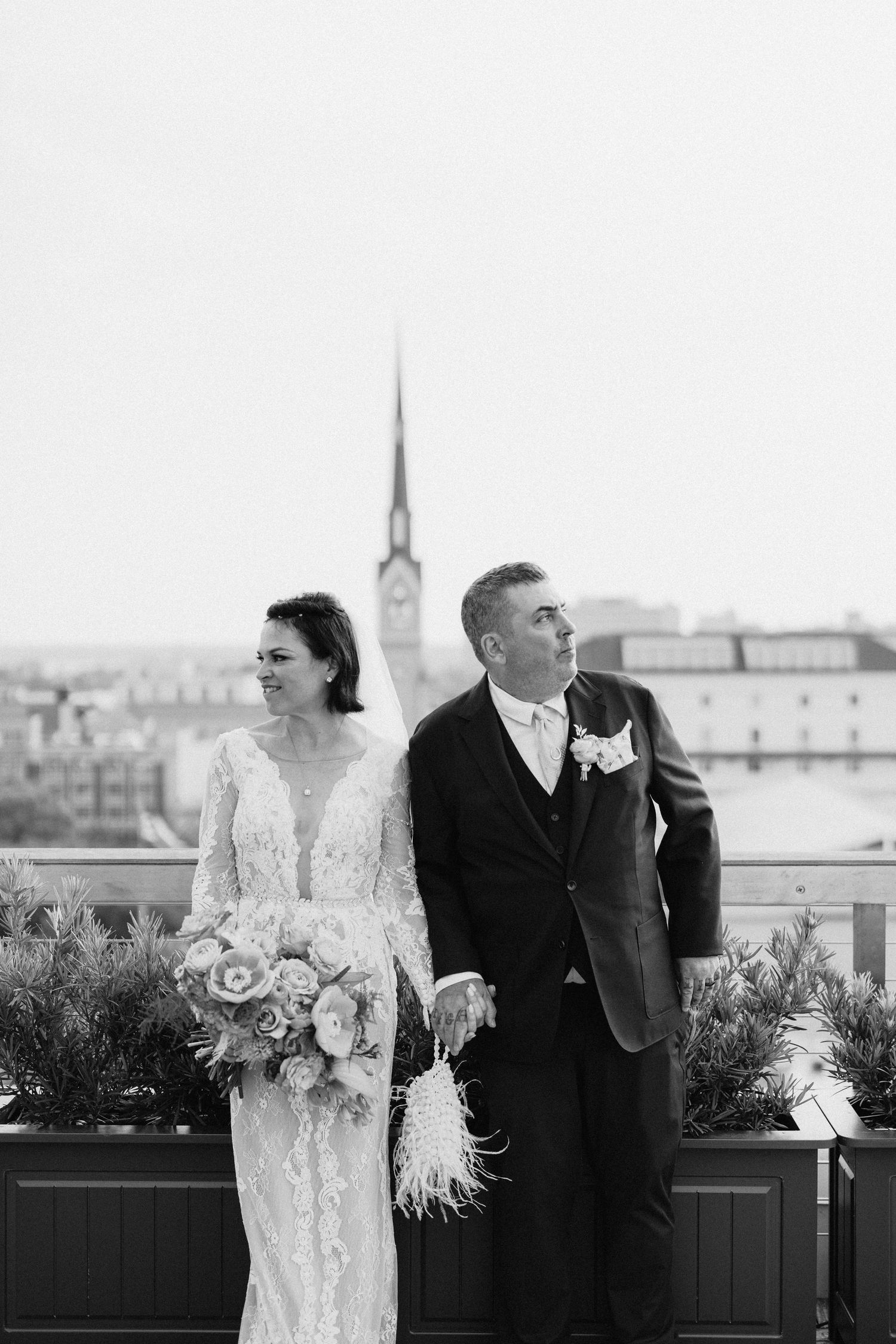 Black and white photo of bride in lace gown and groom in suit posing on rooftop with city skyline backdrop.