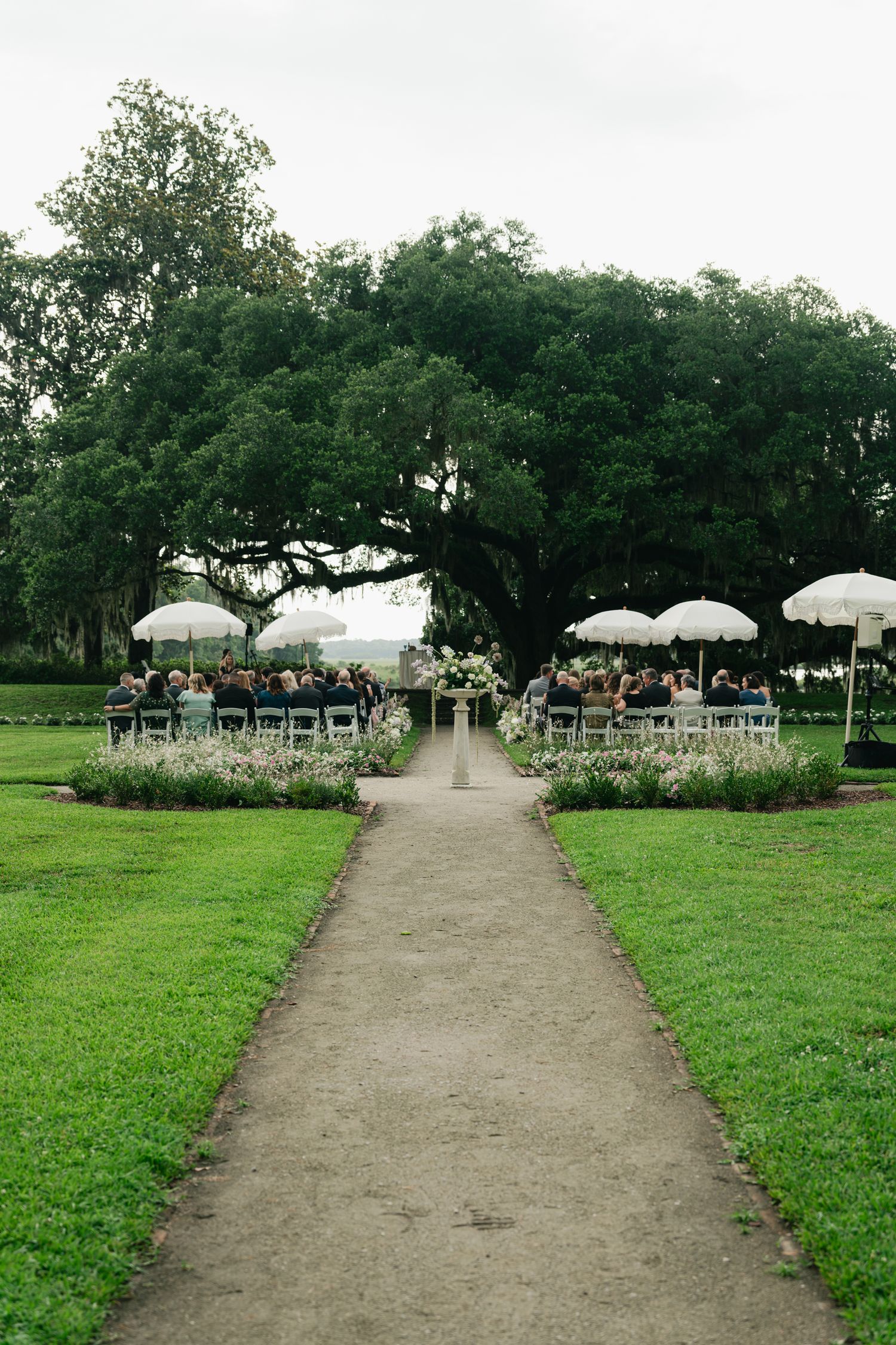 Outdoor wedding ceremony under large oak tree with white umbrellas and floral aisle decorations.