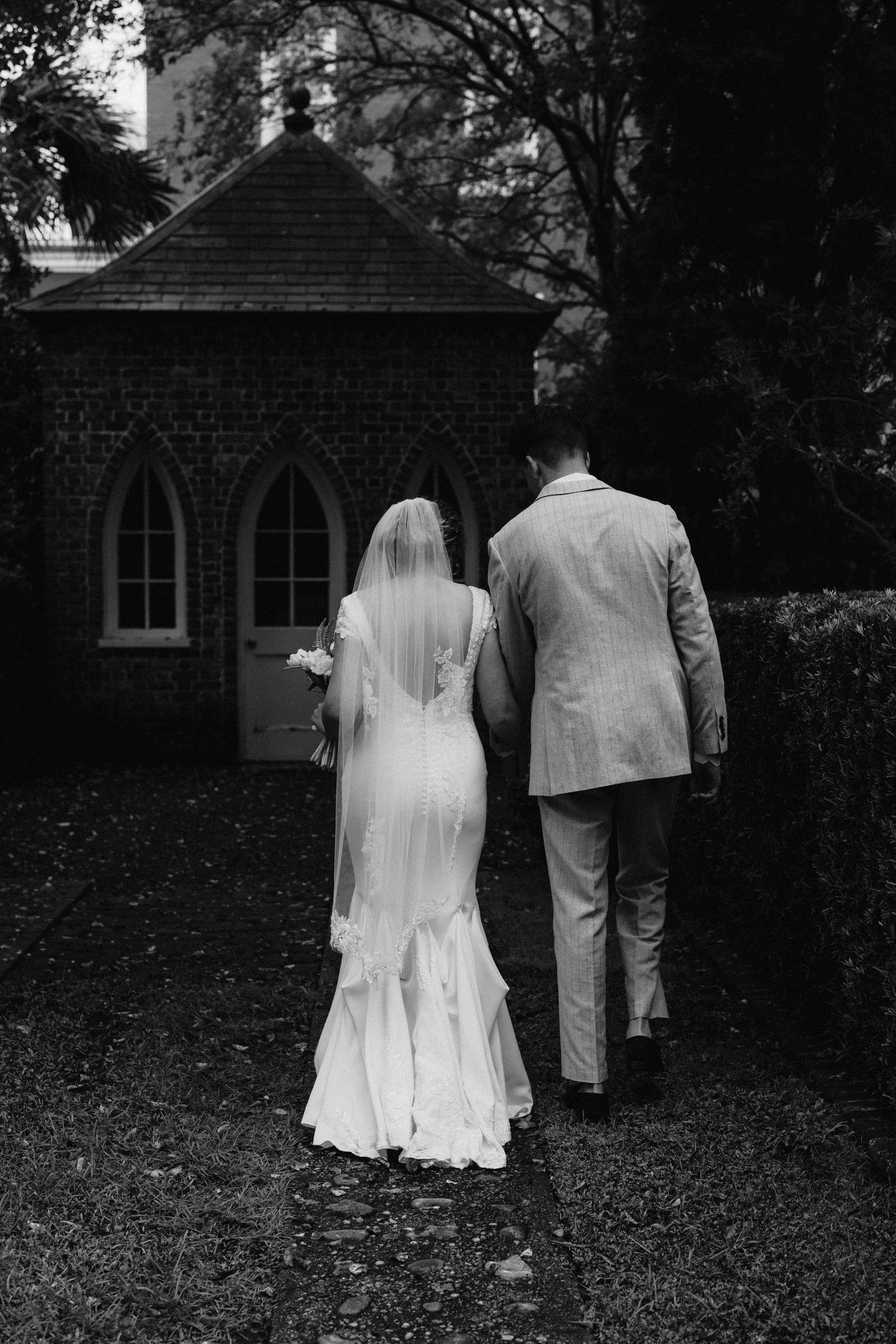 Black and white photo of bride and groom walking toward a historic brick chapel on their wedding day.