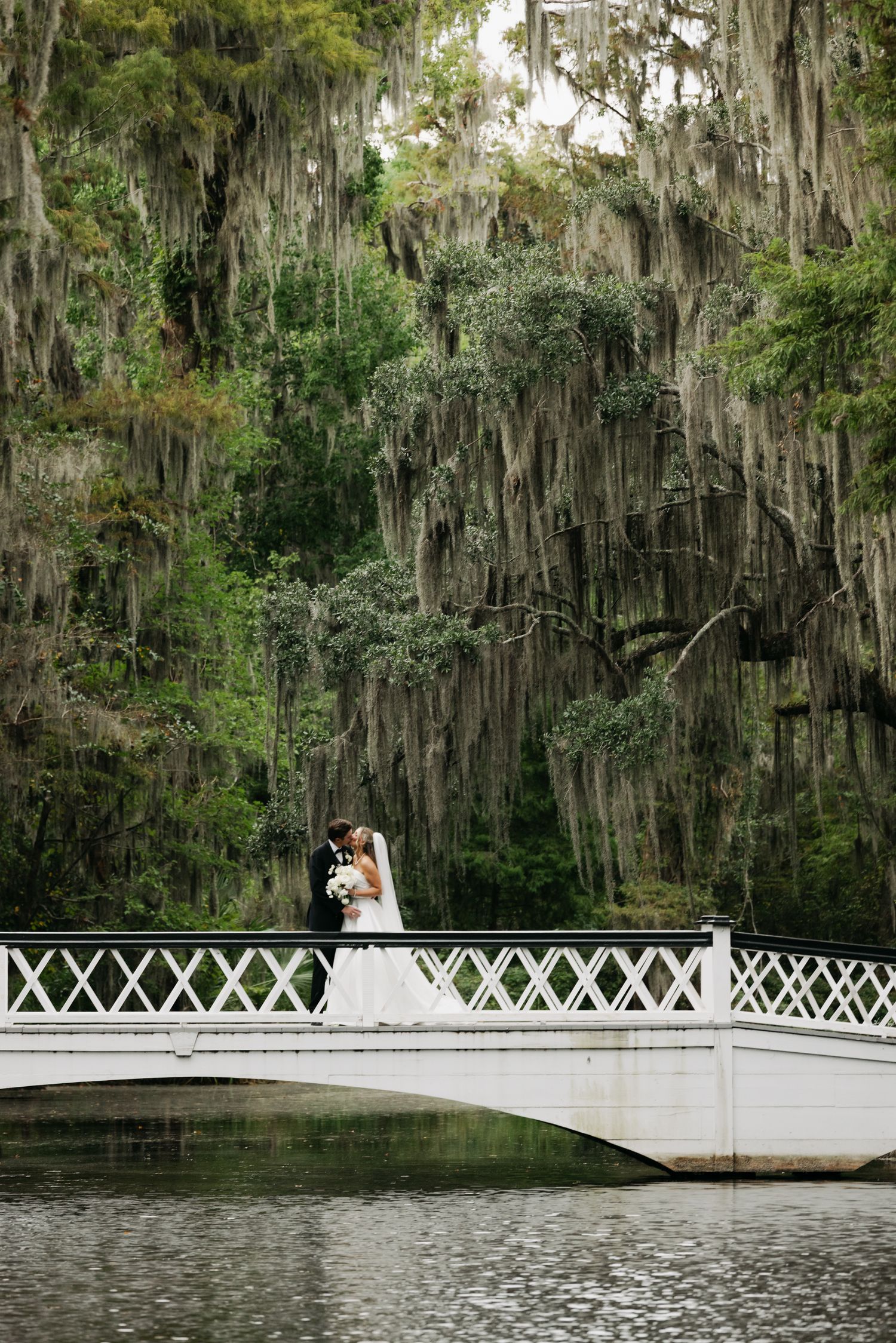 Bride and groom kissing on white bridge over water surrounded by Spanish moss-draped trees.