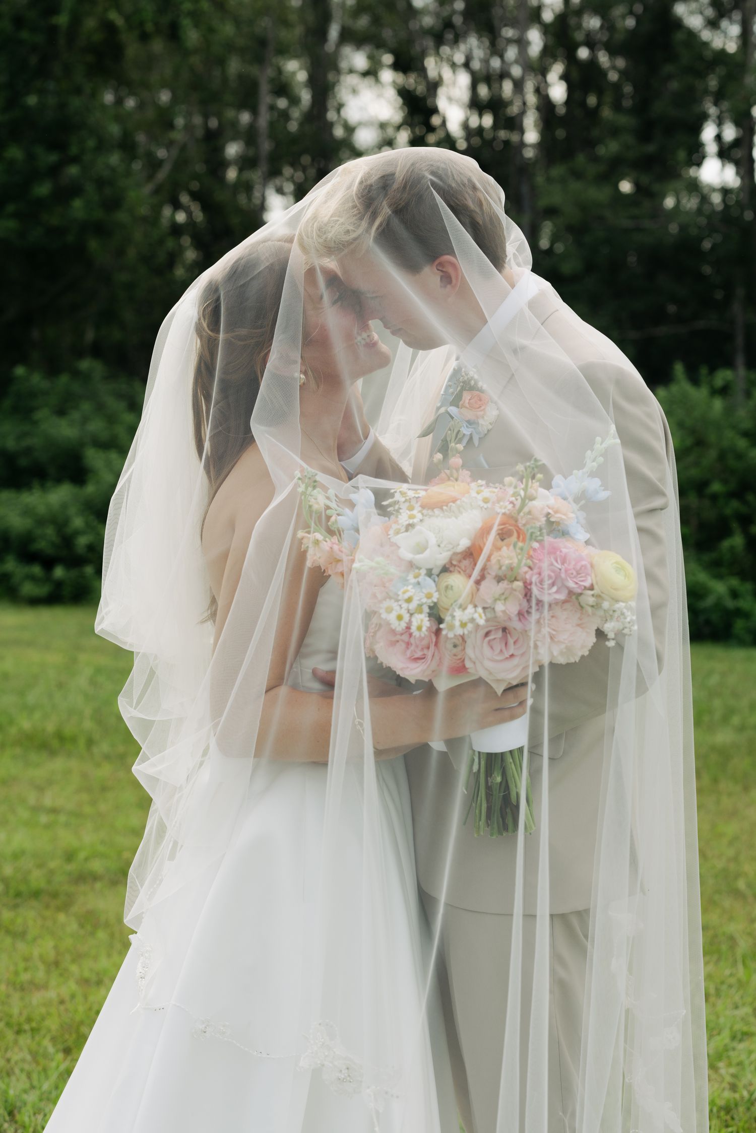 Bride and groom share a kiss hidden beneath a flowing veil, bride holding pastel pink and white bouquet.
