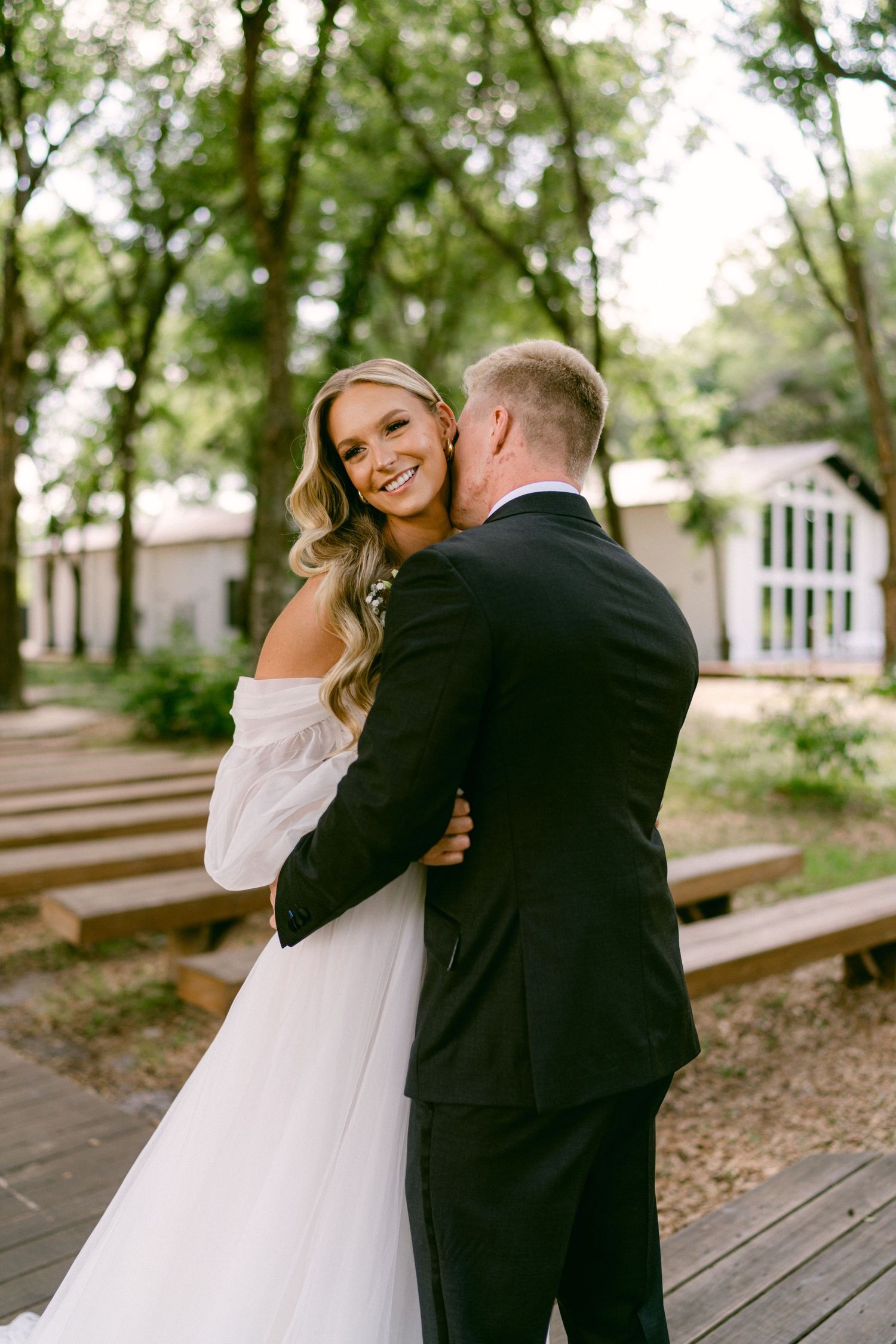 Bride in off-shoulder white gown smiles over shoulder as groom in black suit embraces her outdoors among trees.