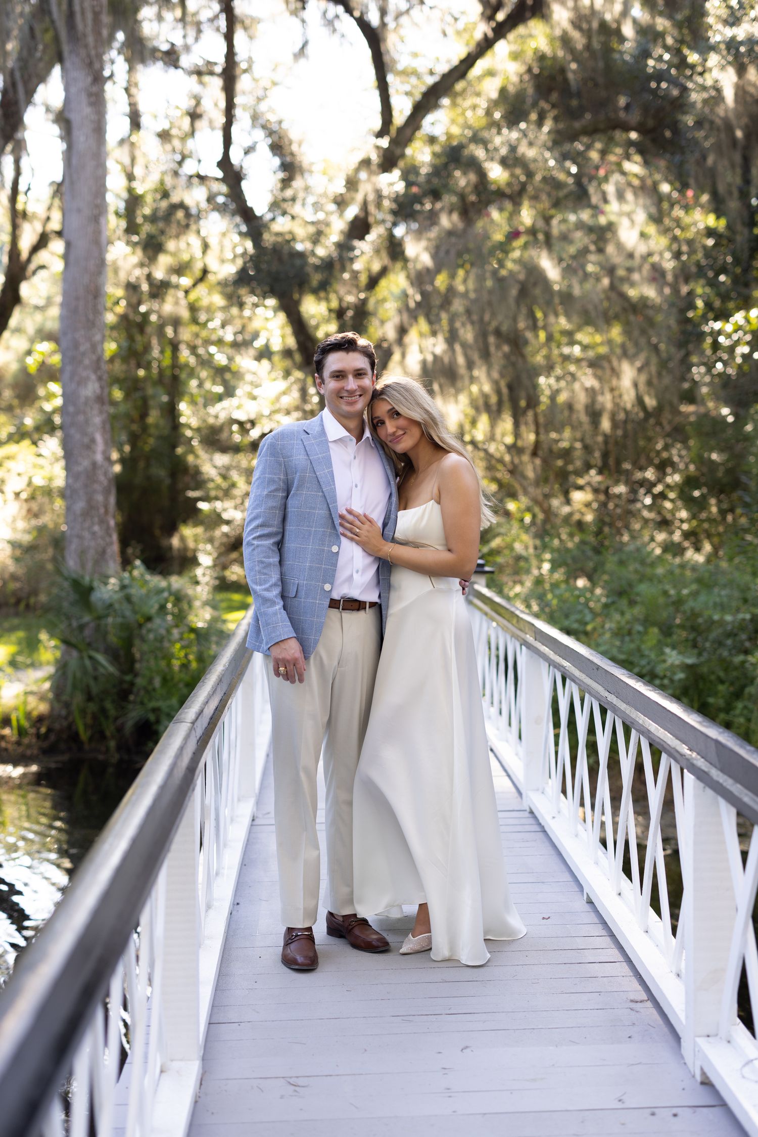 Couple in elegant attire embracing on a white bridge surrounded by lush greenery and Spanish moss-draped trees.