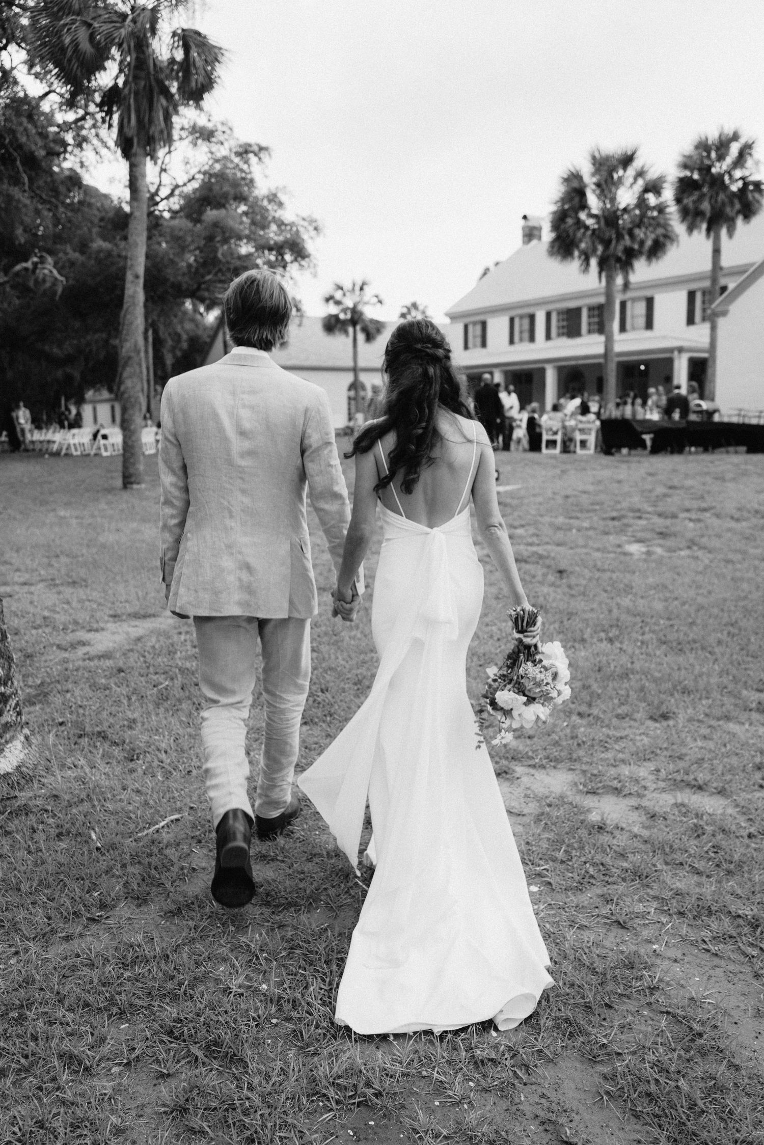 Black and white photo of bride and groom walking hand in hand toward a Southern estate surrounded by palm trees.