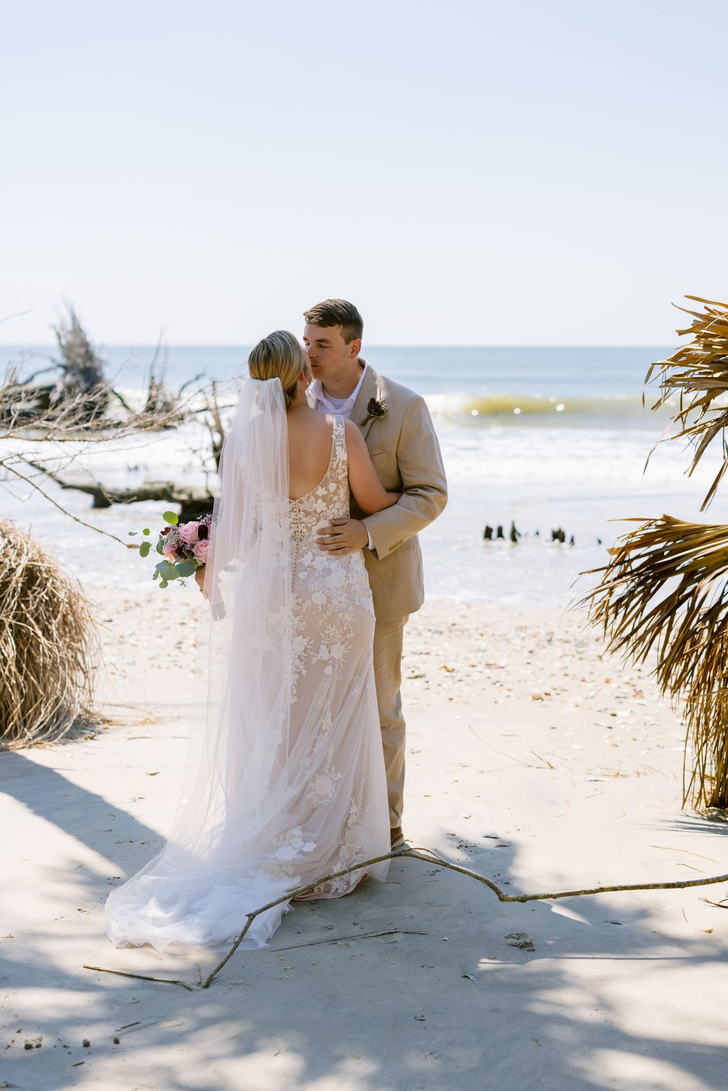 Bride and groom share first kiss on sandy beach wedding ceremony with ocean waves and palm fronds in background.