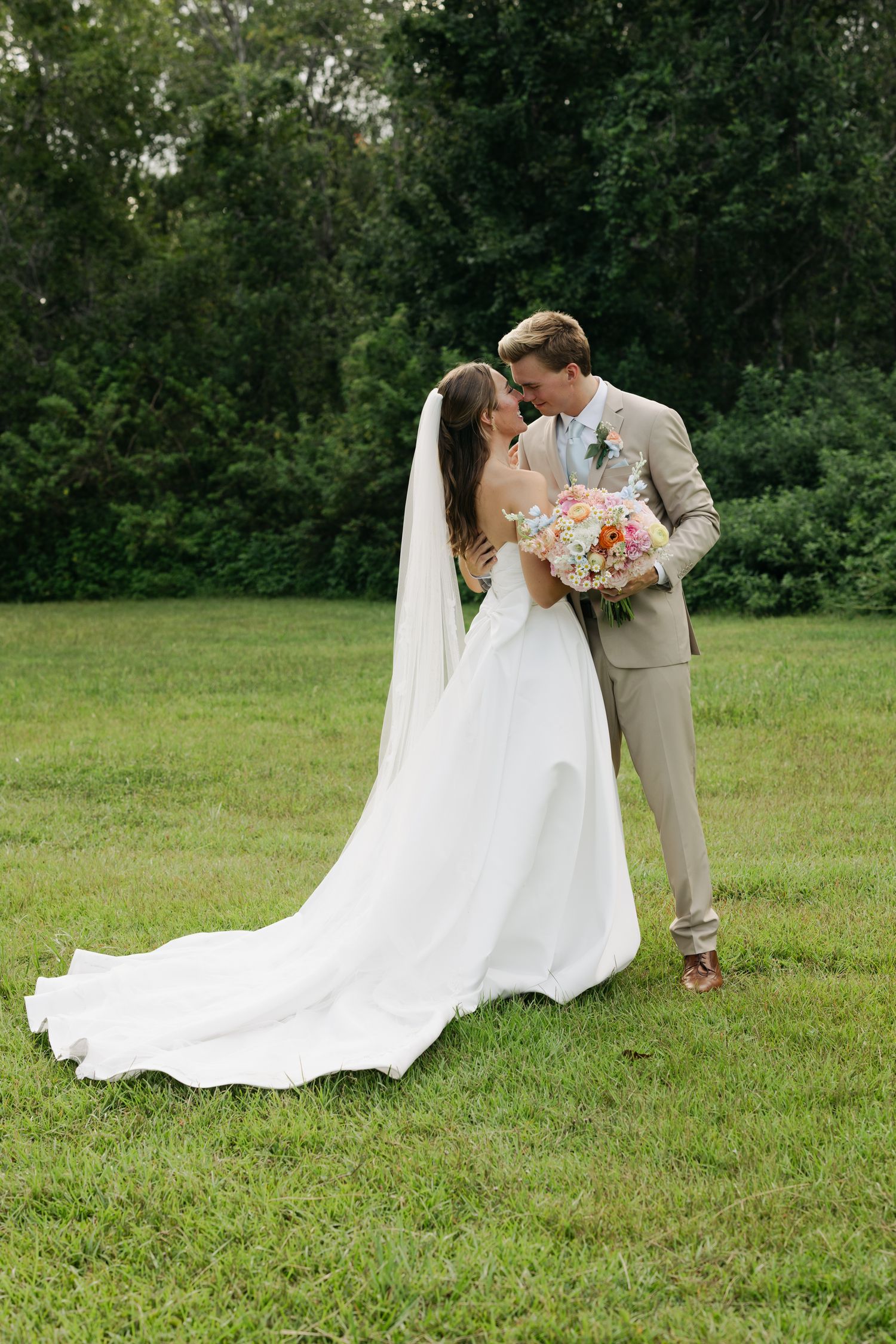 Bride in white gown with veil and groom in tan suit share a romantic moment on green lawn with trees.