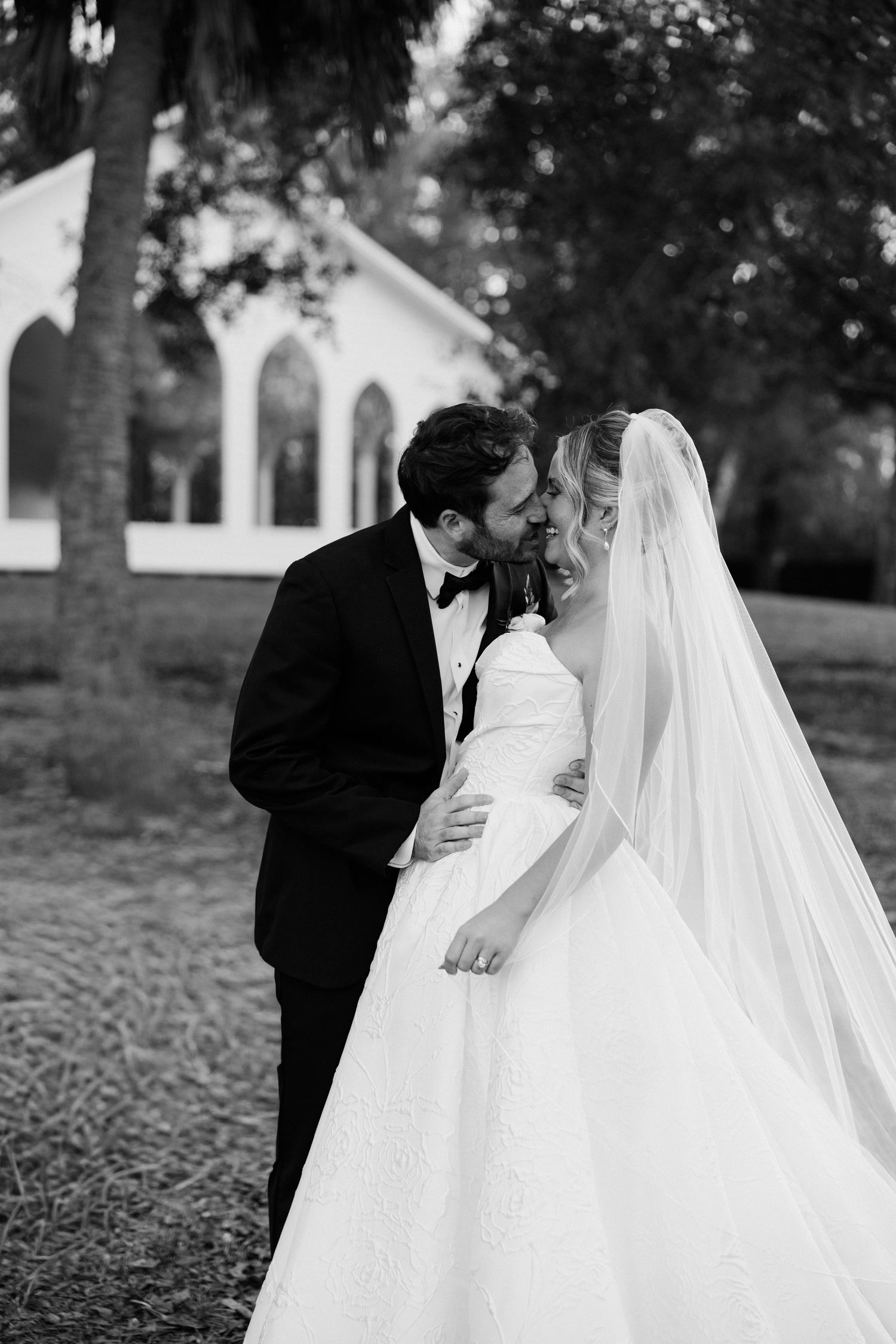 Black and white photo of a bride and groom sharing a romantic moment outside a white chapel.
