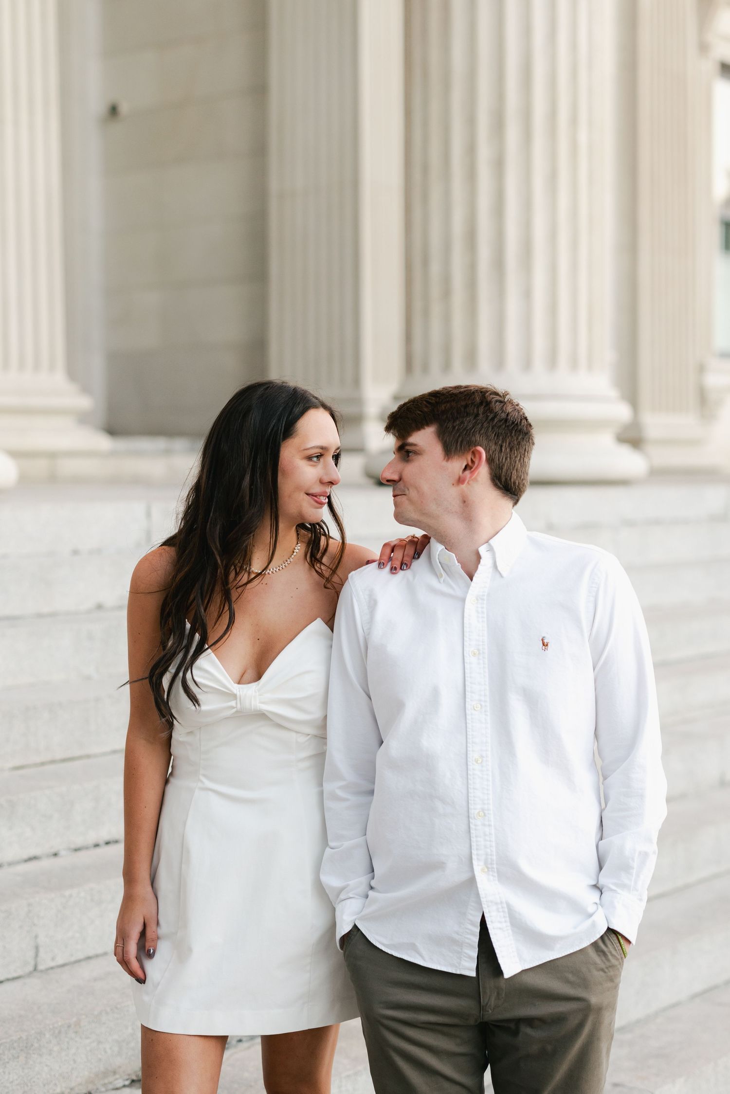 A couple in white outfits share a romantic glance on stone steps with classical columns behind them.