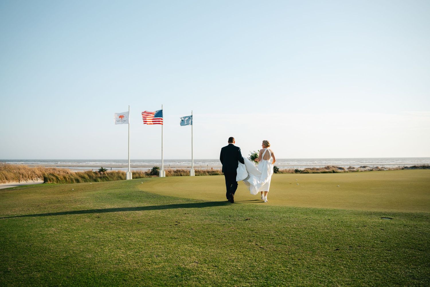 Bride and groom walking on golf course with ocean view and flags in background on wedding day.