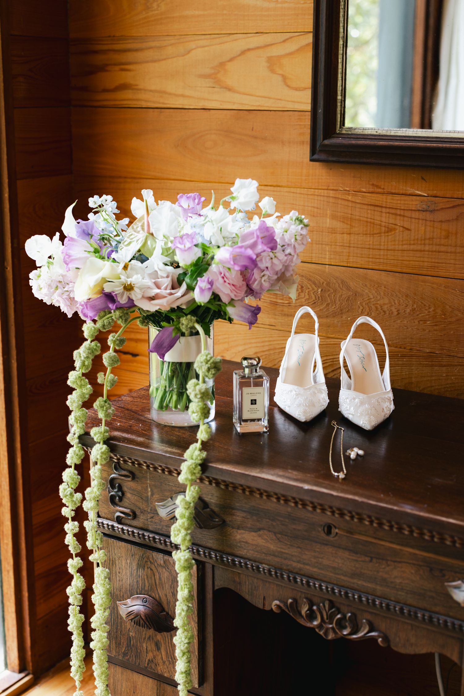 Elegant bridal flat lay with purple floral bouquet, white lace heels, Jo Malone perfume, and jewelry on antique wooden dresser.