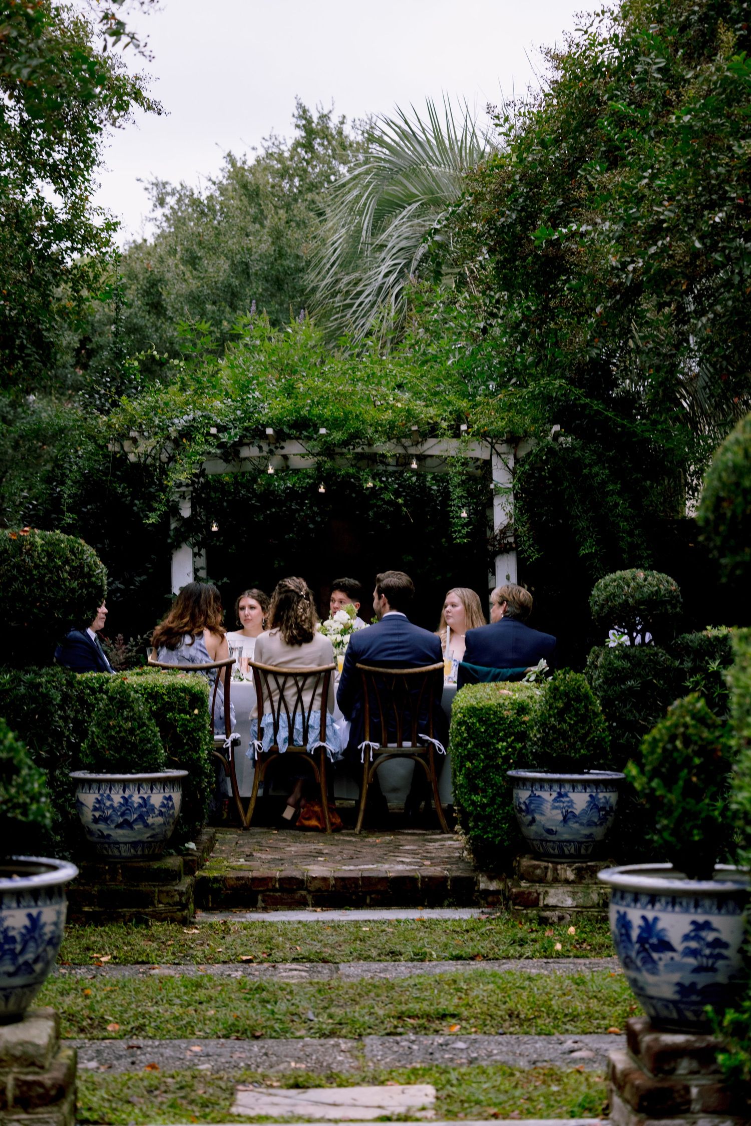 Elegant outdoor garden dinner party with guests seated under pergola surrounded by topiaries.