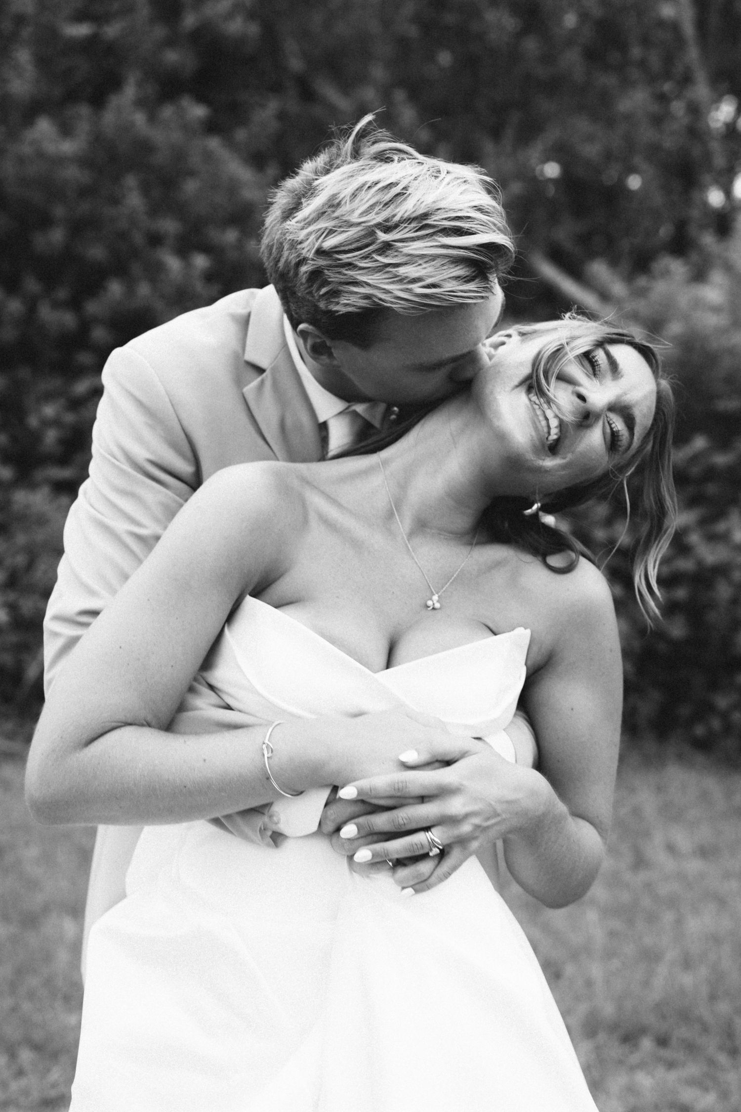 Groom kissing laughing bride on the neck while embracing her from behind in black and white wedding photo.