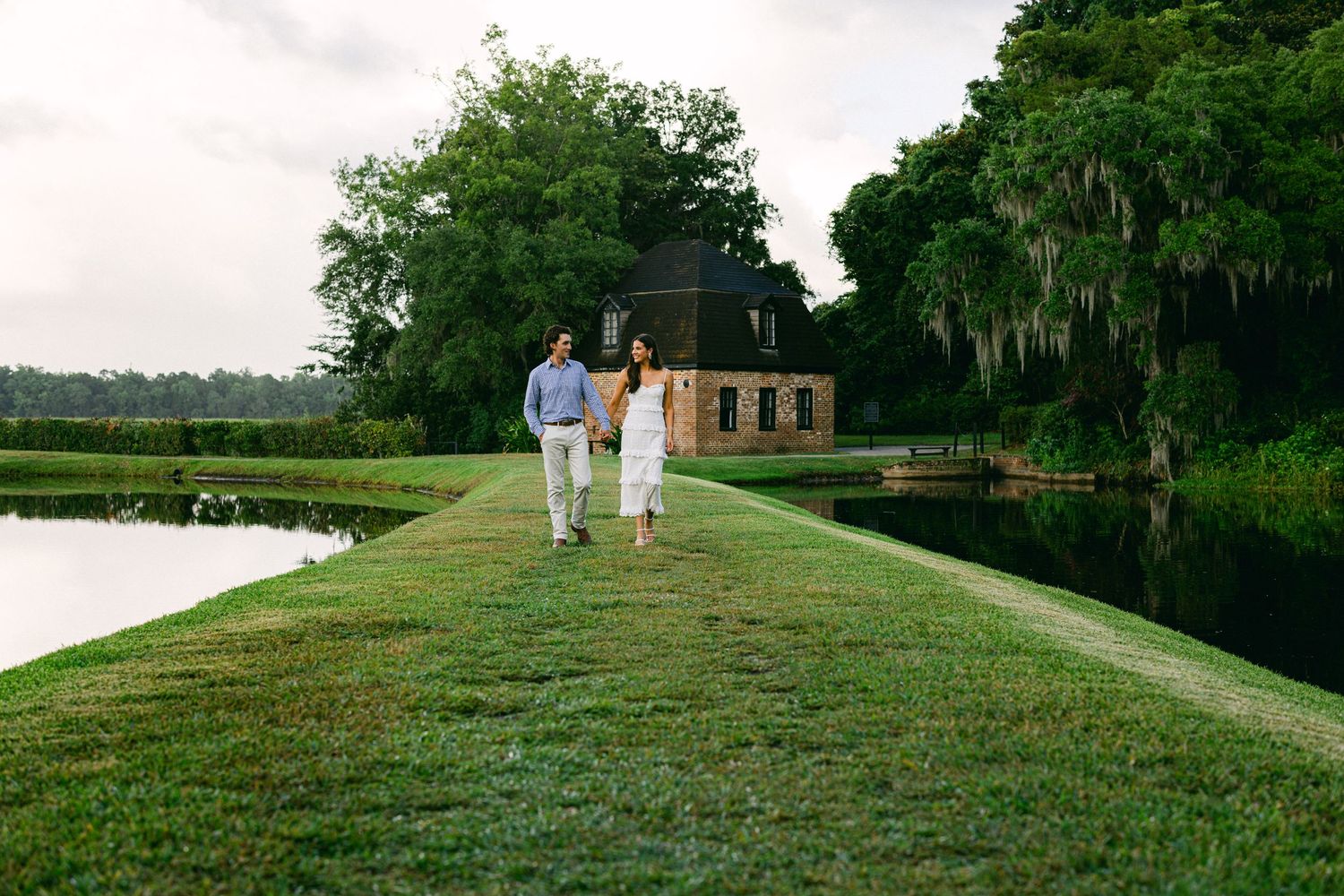 Couple walking hand-in-hand along a lush green lawn beside a serene pond at a historic plantation.