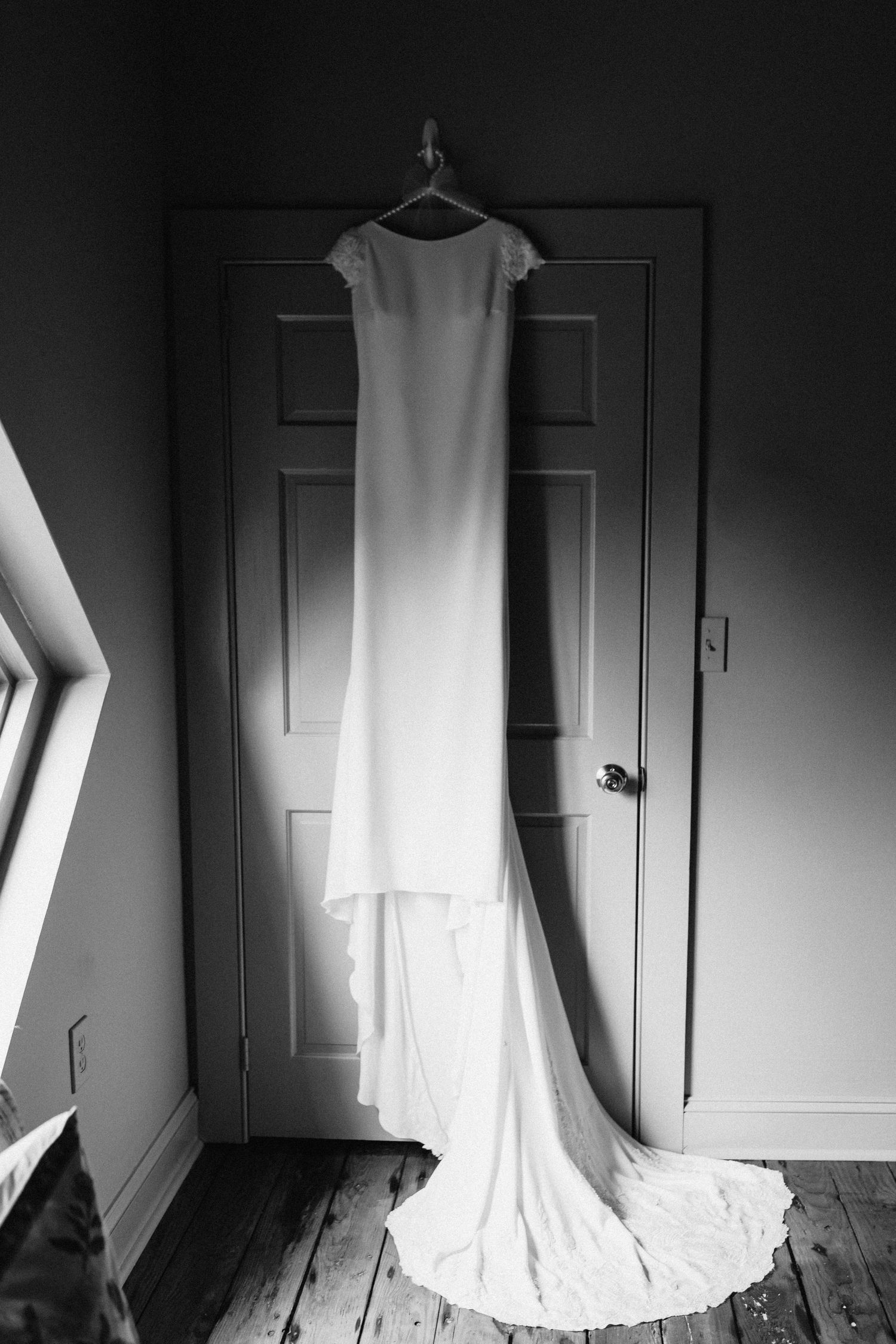 Elegant white wedding dress with long train hanging on door hanger in black and white photo.