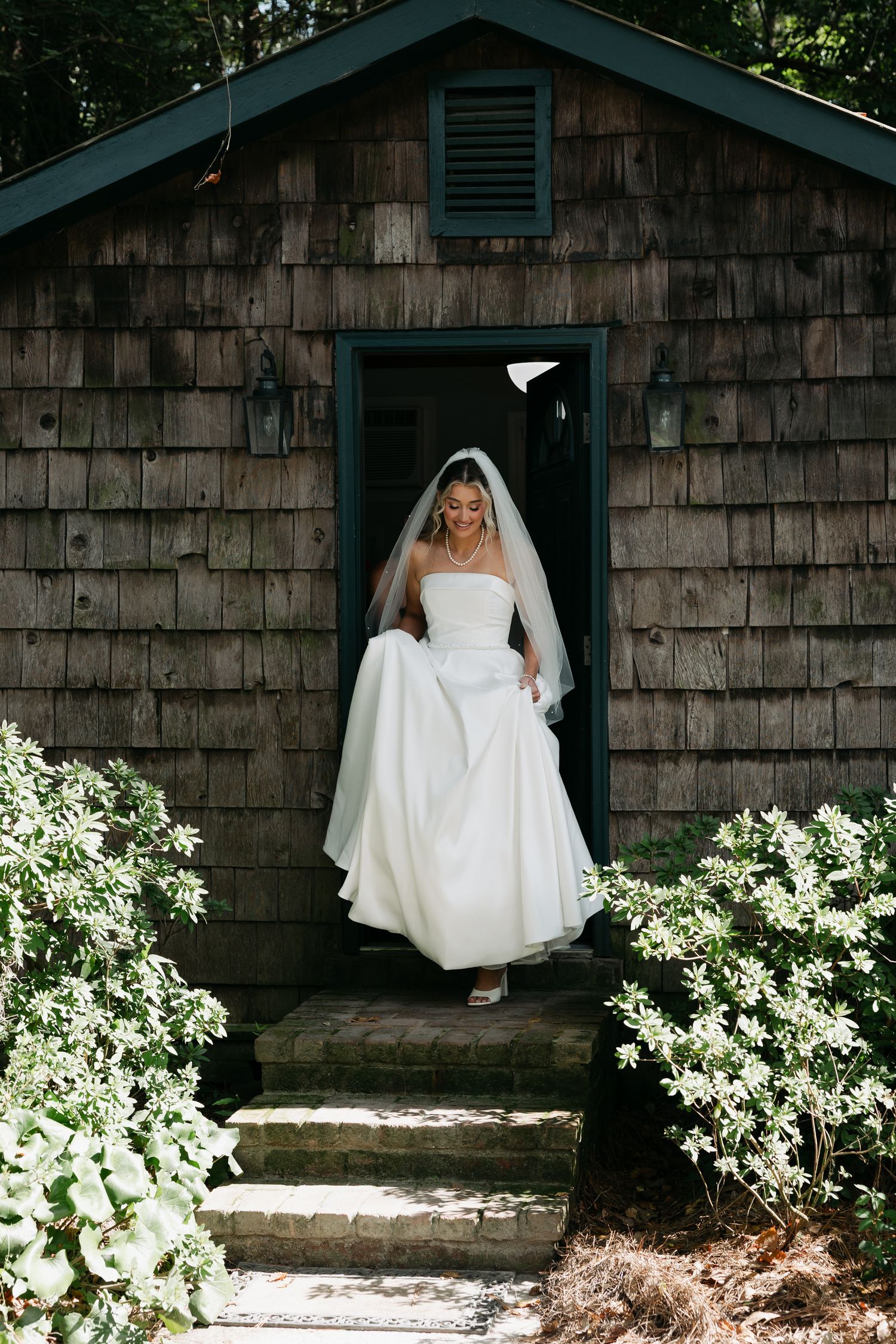 Radiant bride in classic white strapless gown and veil steps out of rustic shingle cottage doorway surrounded by greenery.