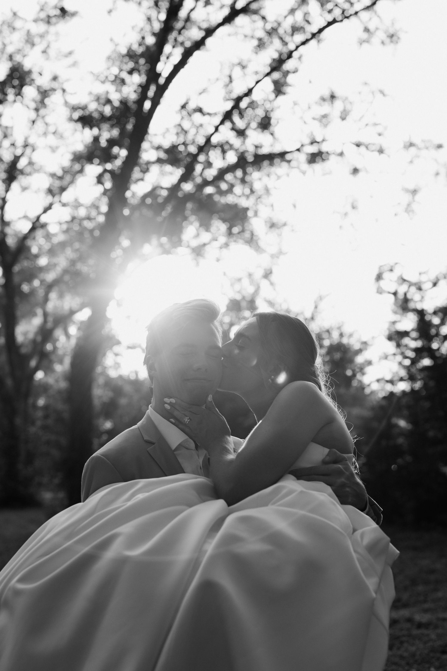 Black and white photo of groom carrying bride in wedding dress, sharing a kiss in sunlit park setting.