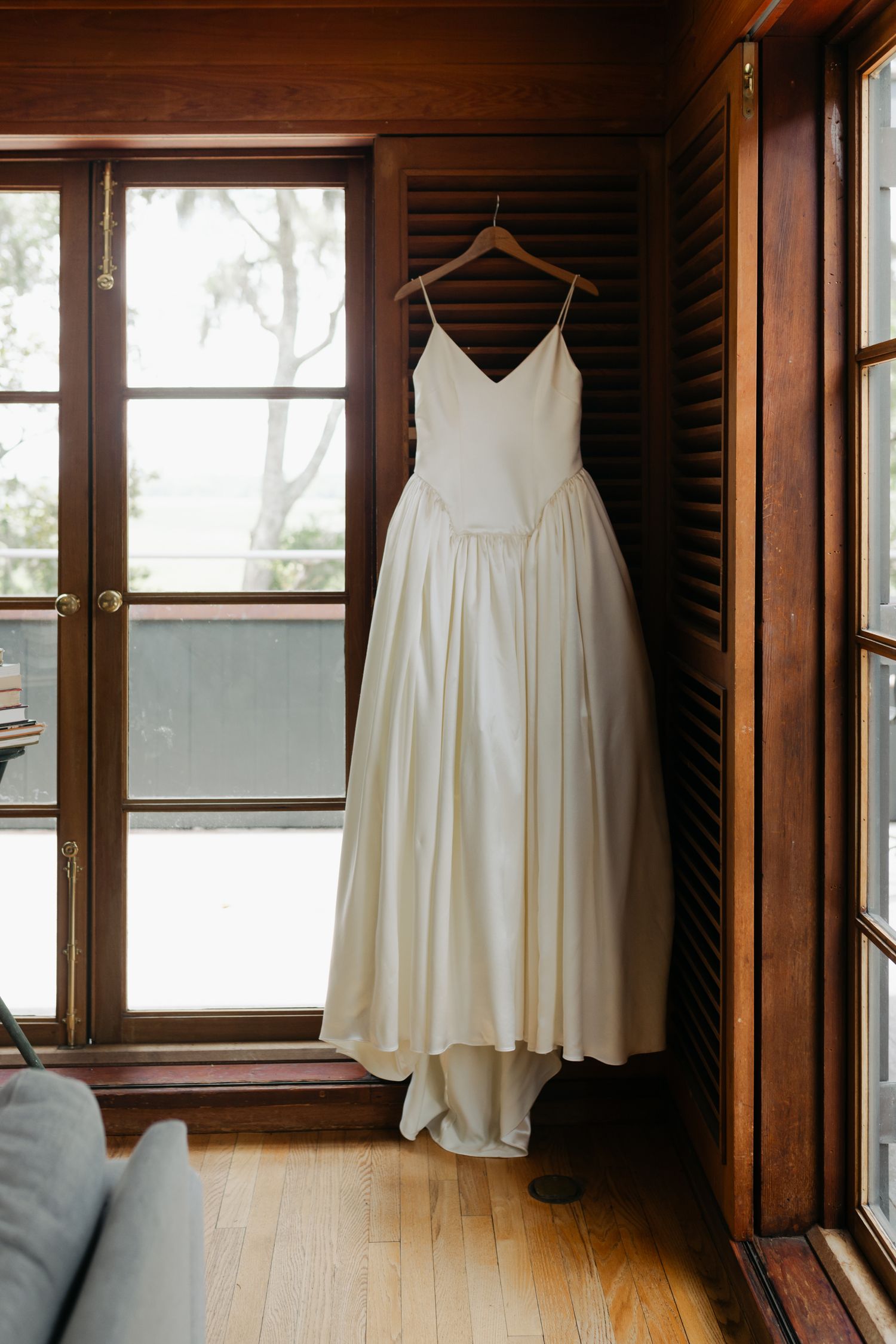 Elegant white satin wedding dress with spaghetti straps hanging on wooden hanger in rustic wood-paneled room.