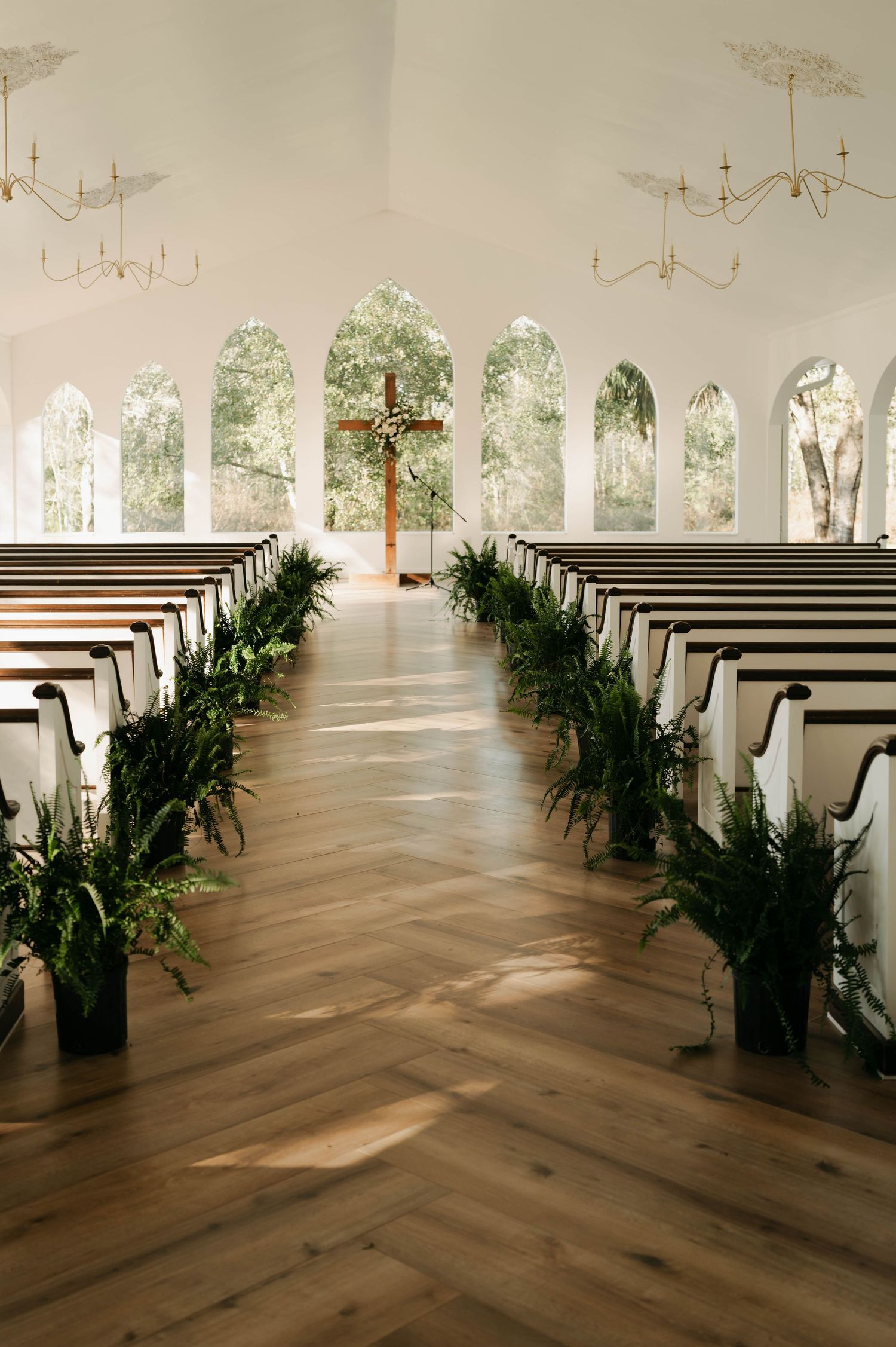 Elegant white chapel with wooden pews, herringbone floor, greenery aisle decor, arched windows, and a wooden cross at the altar.