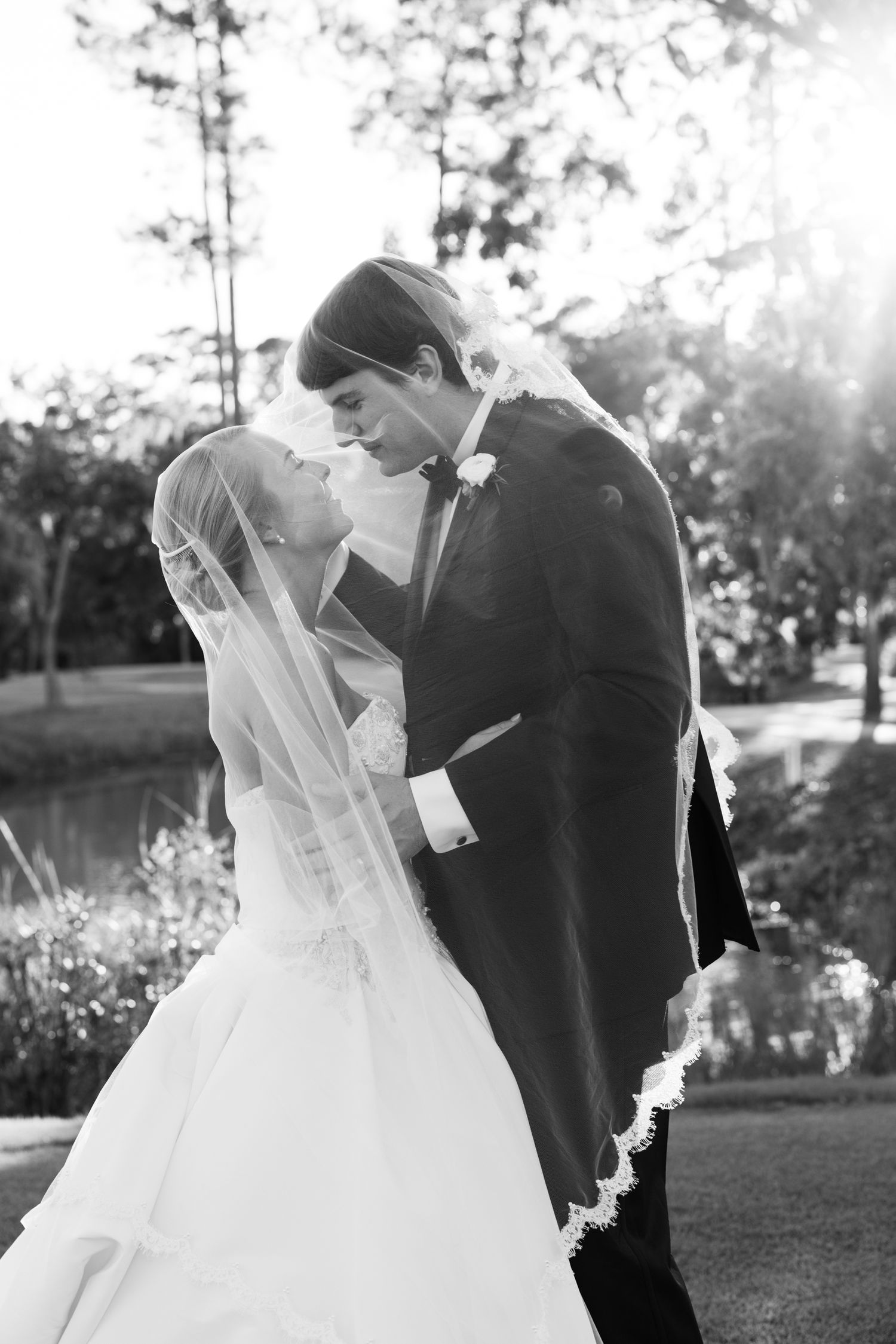 Black and white photo of a bride and groom sharing a romantic moment under her veil outdoors.